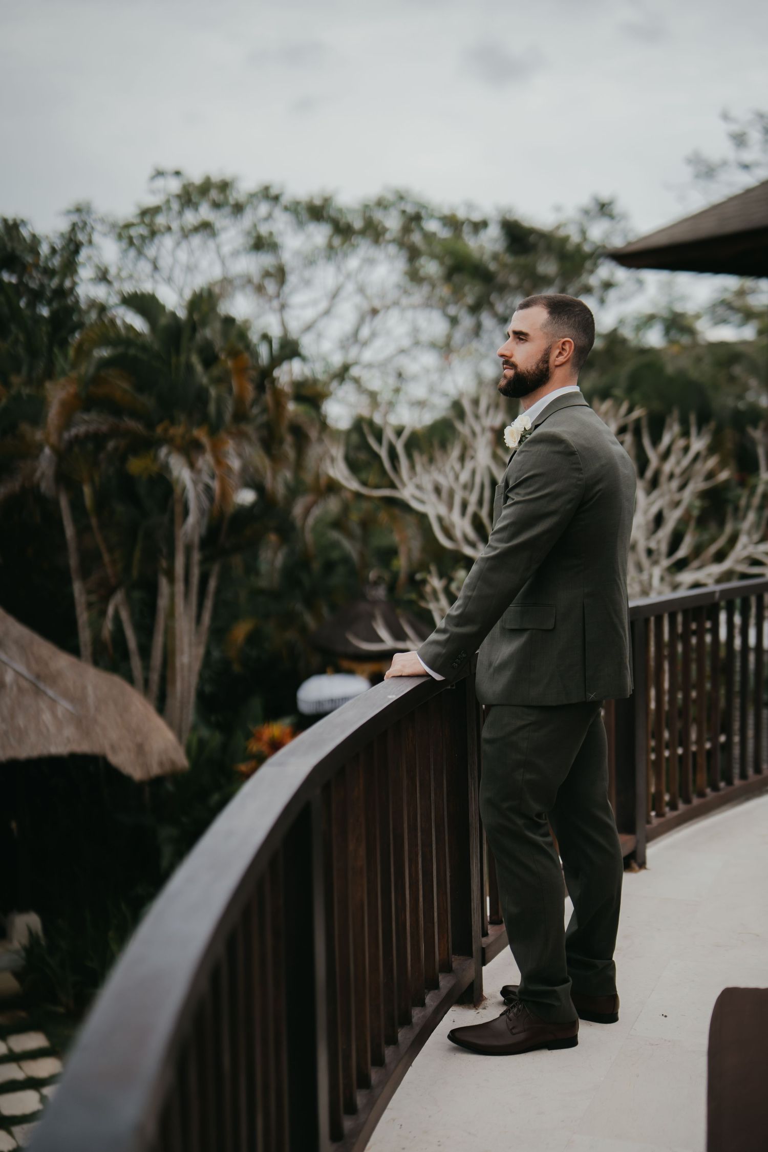 A groom in a dark suit gazes over a wooden balcony railing surrounded by lush tropical trees.