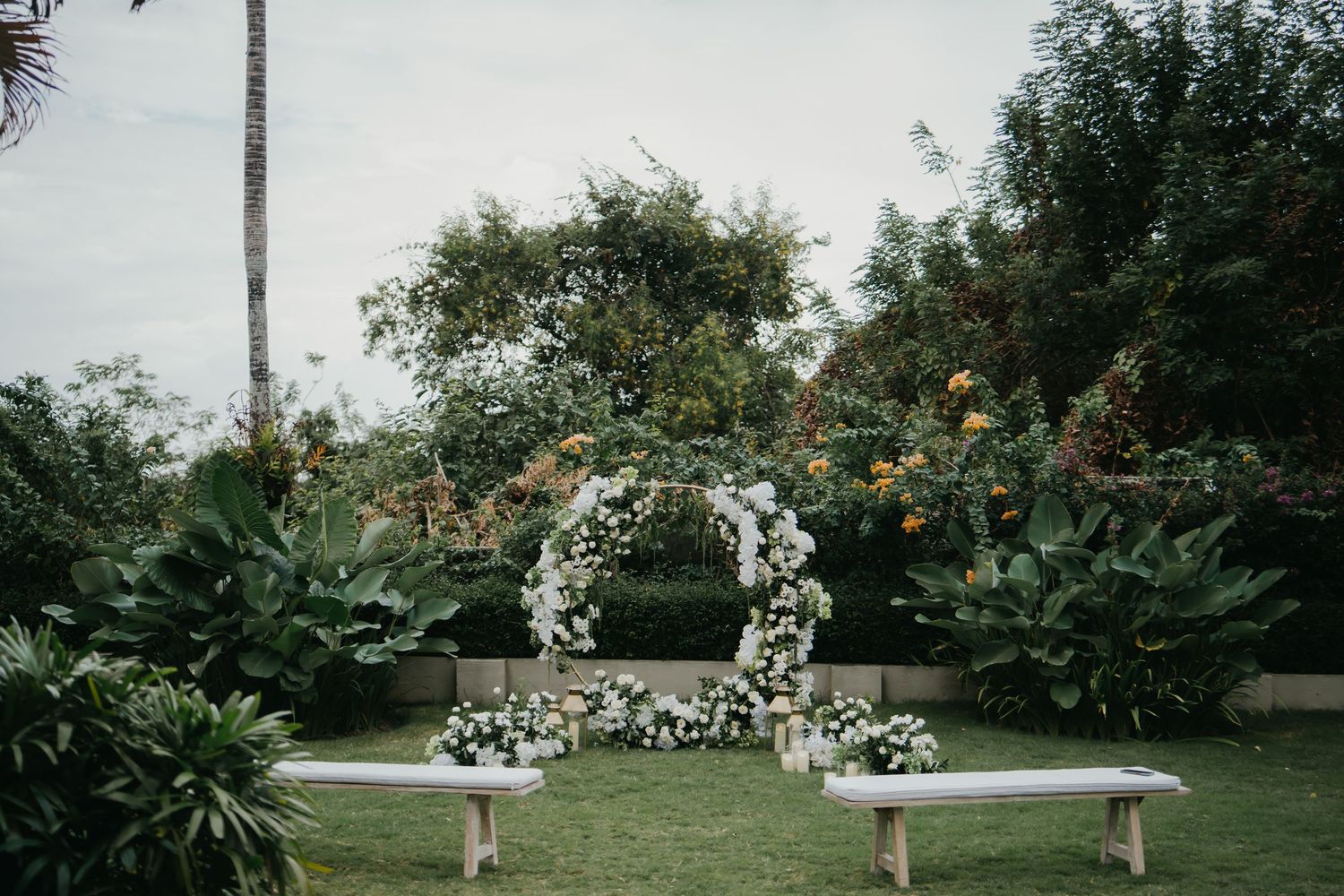 Circular white floral arch with wooden benches on green lawn for intimate outdoor garden wedding ceremony.