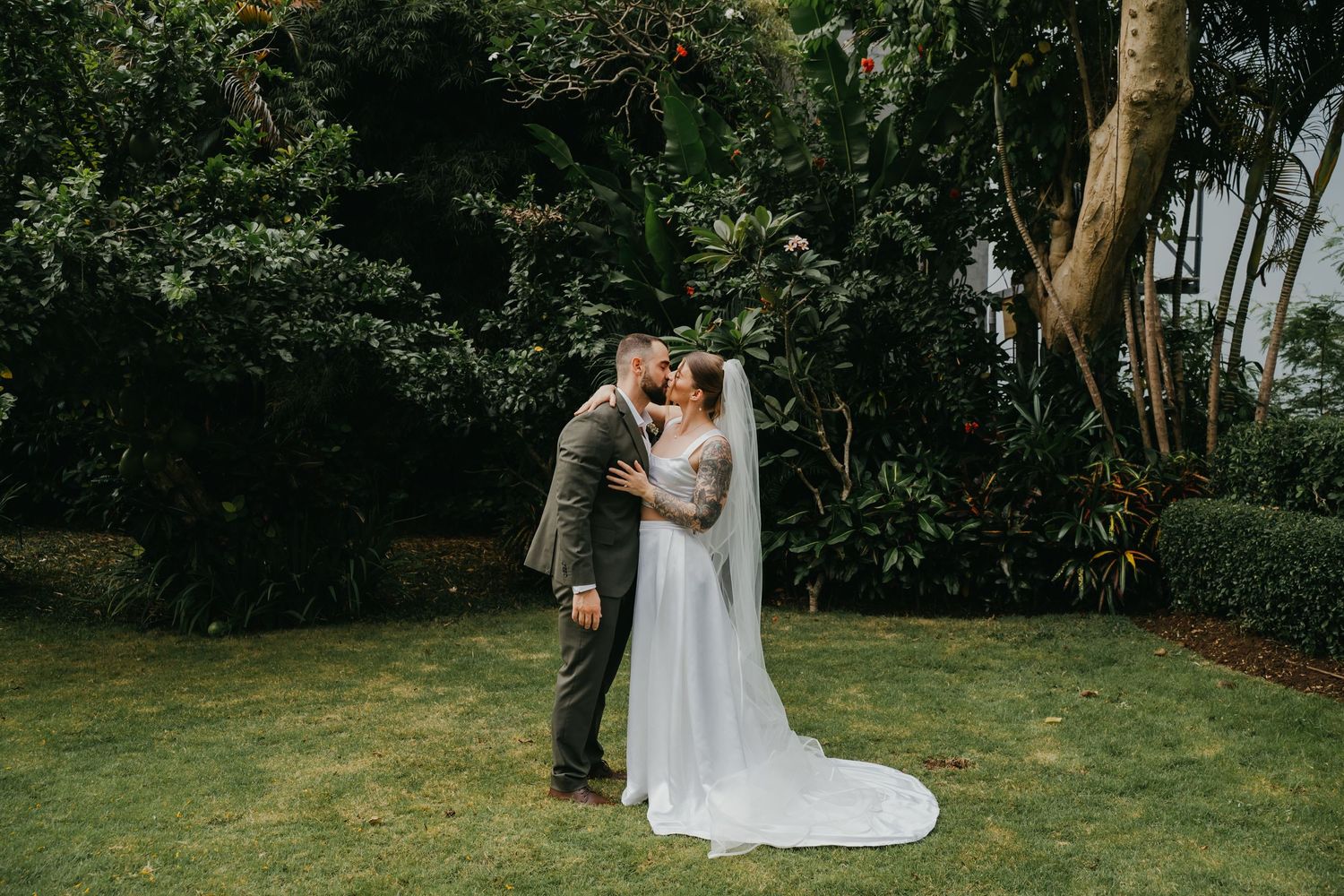 Newlyweds share romantic kiss on lush green lawn surrounded by tropical garden trees.