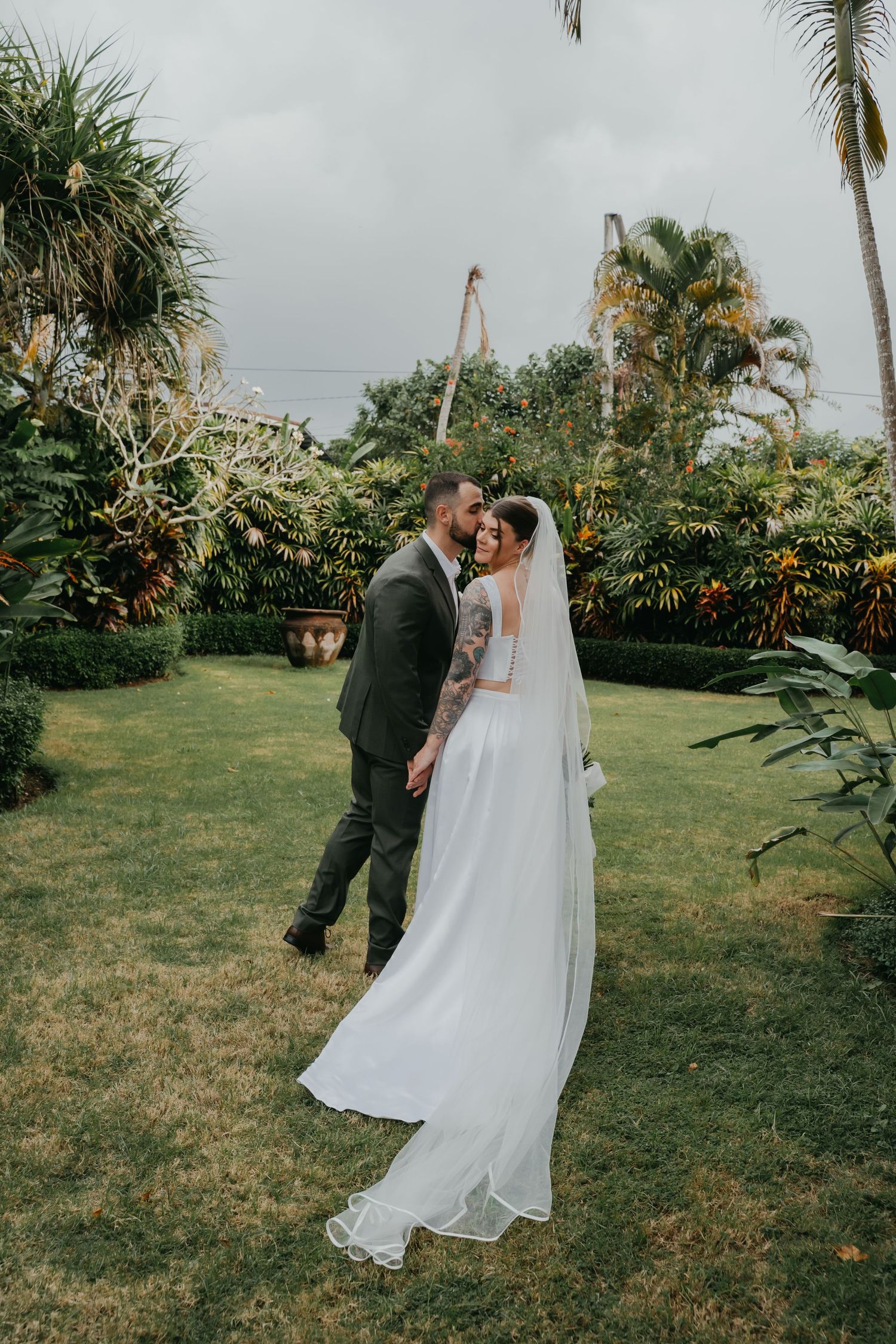 Bride and groom sharing intimate moment in lush tropical garden, brides veil trailing on grass.