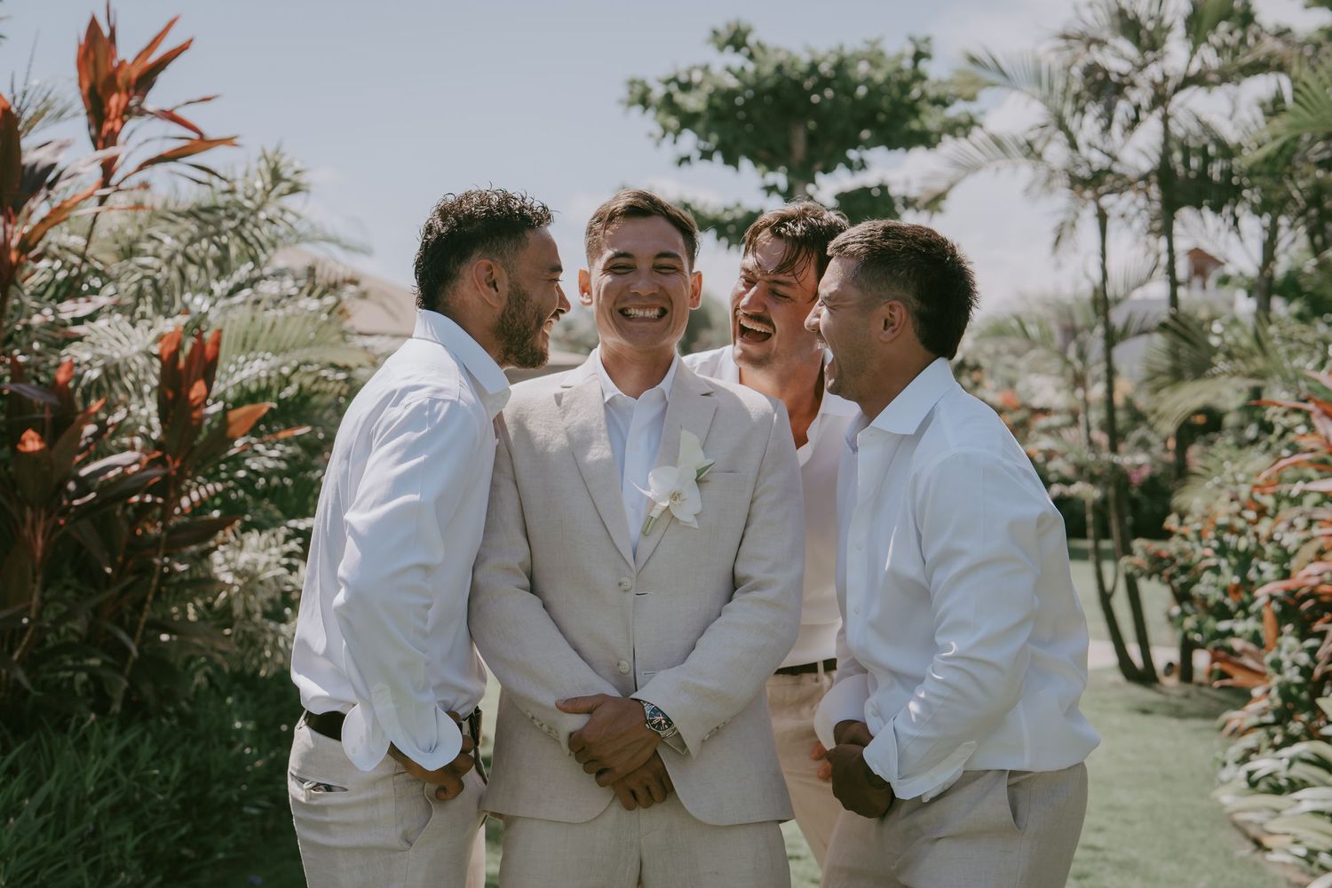 Groomsmen in light colored suits share a candid moment together in a tropical garden setting.