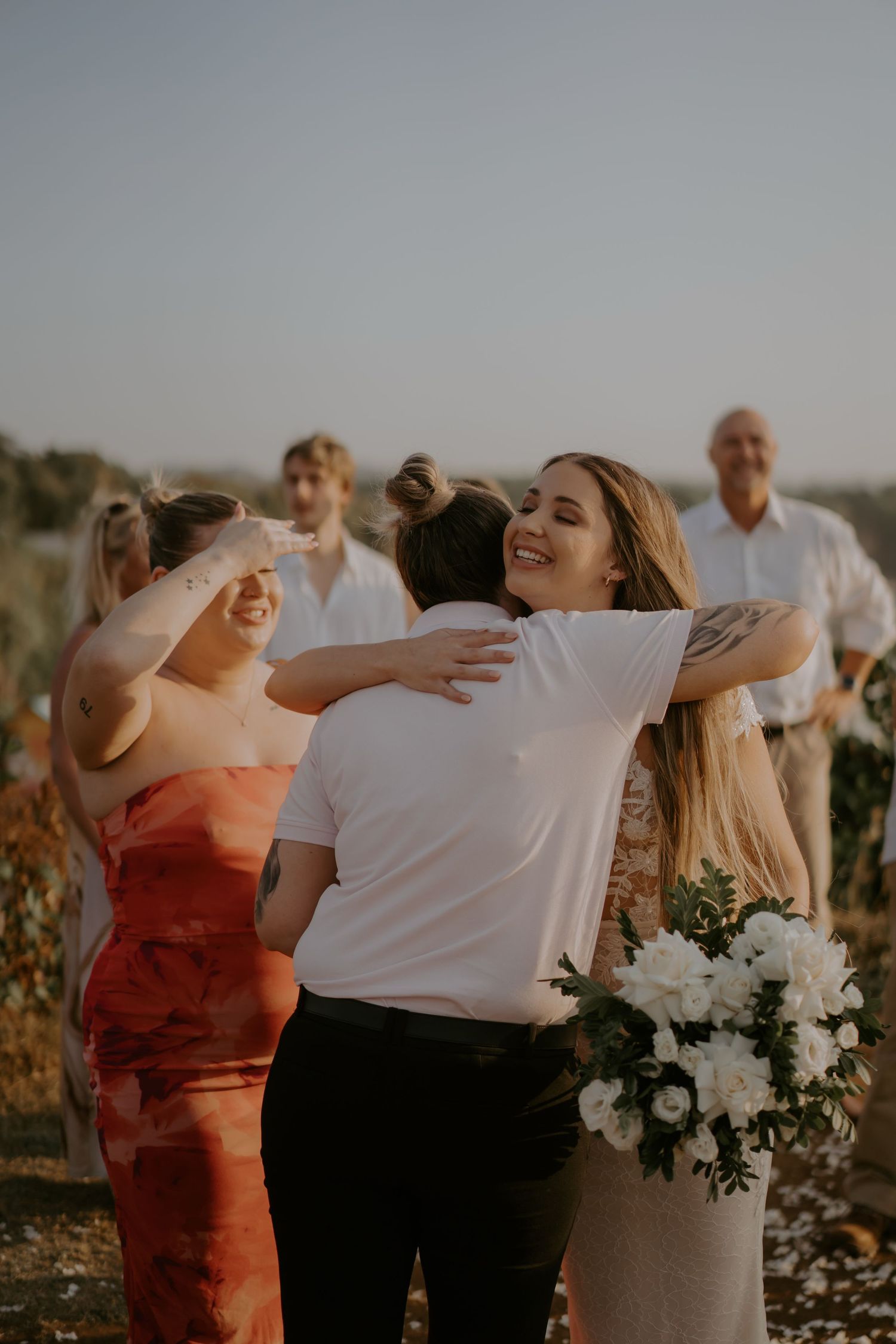 A group of wedding guests dancing and celebrating together in golden sunset light with white floral bouquets.