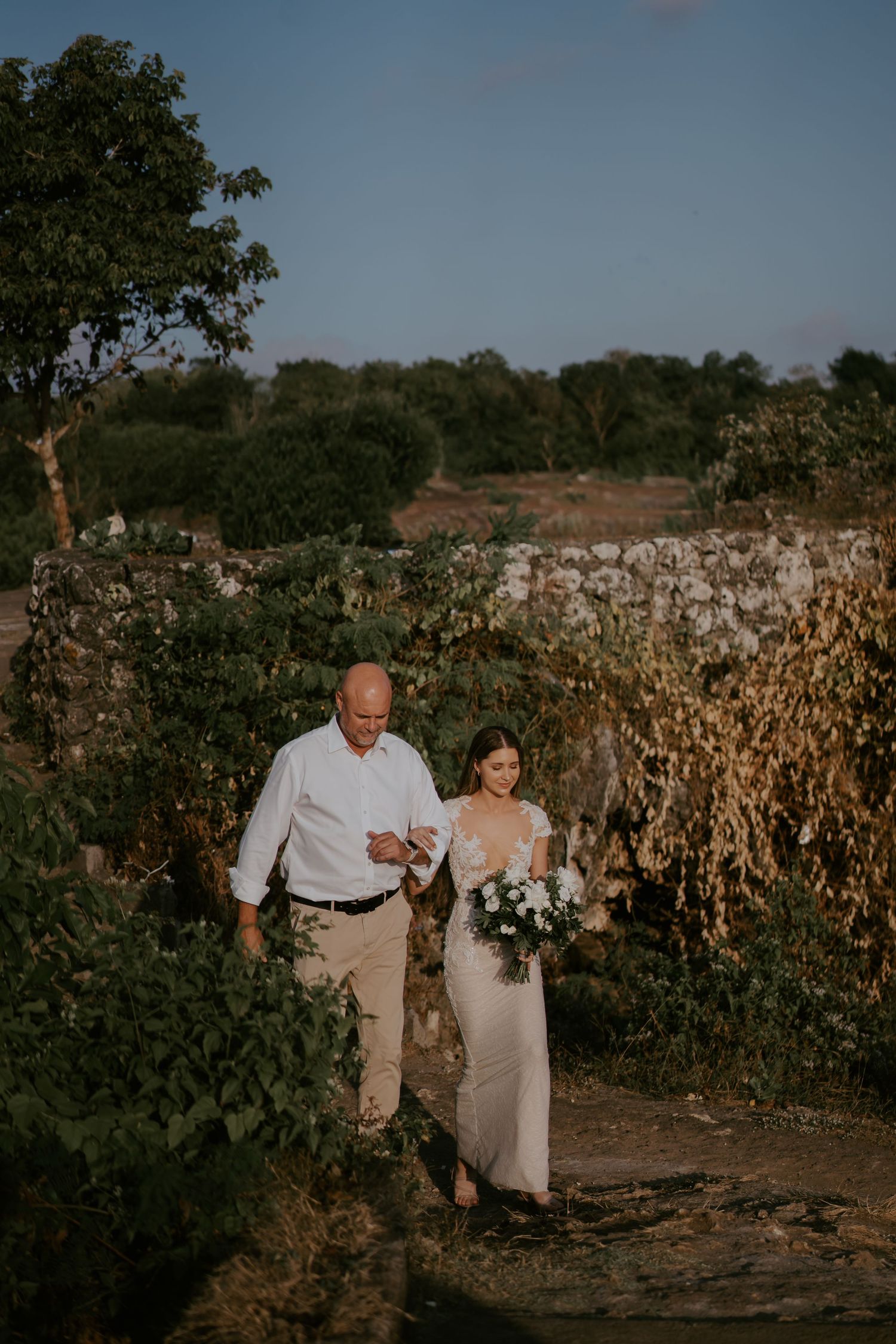 A couple walks along a garden path during a sunset wedding ceremony in a rustic outdoor setting.