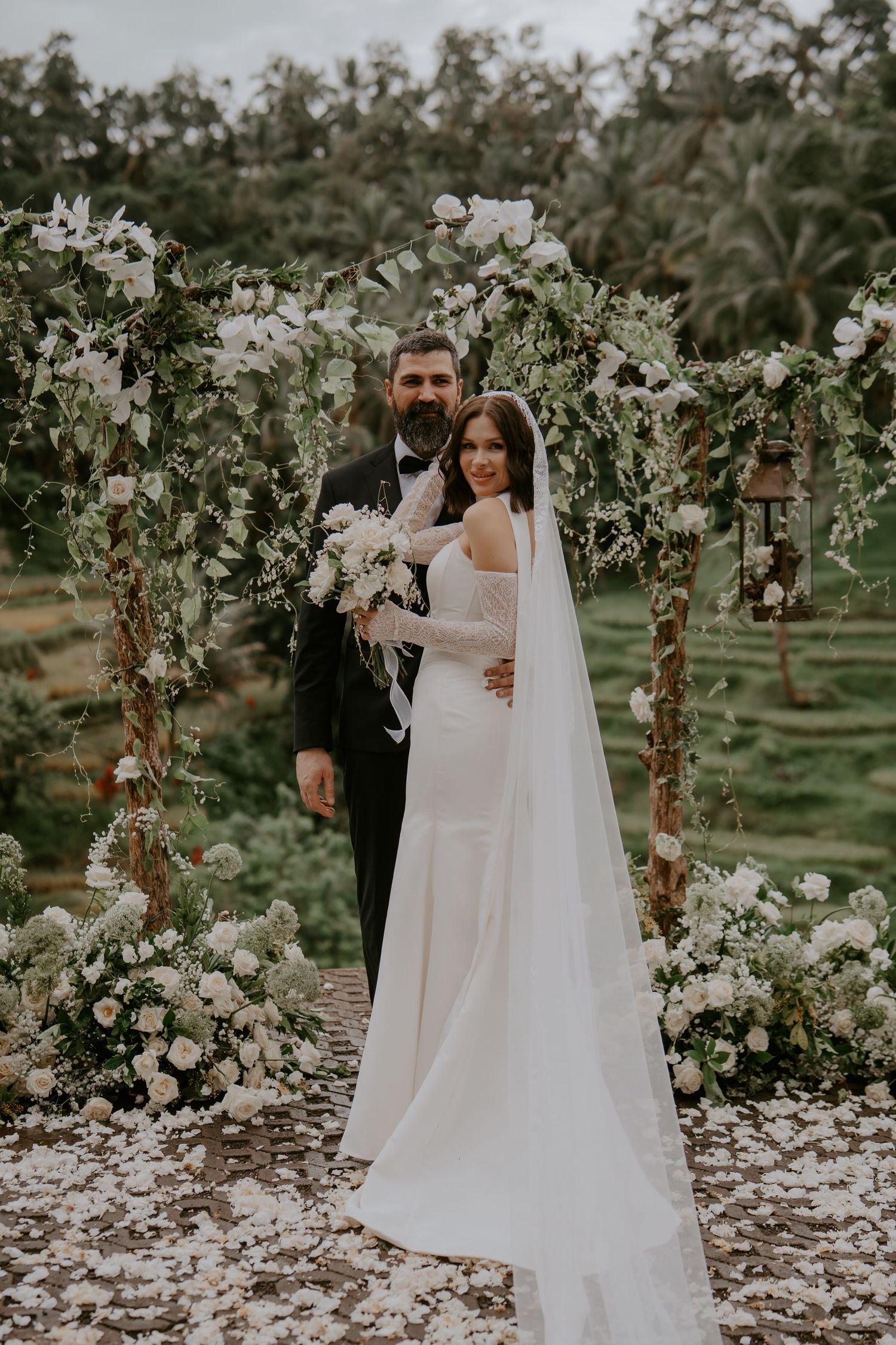 A couple poses in a romantic garden setting surrounded by white floral arrangements and trees on their wedding day.