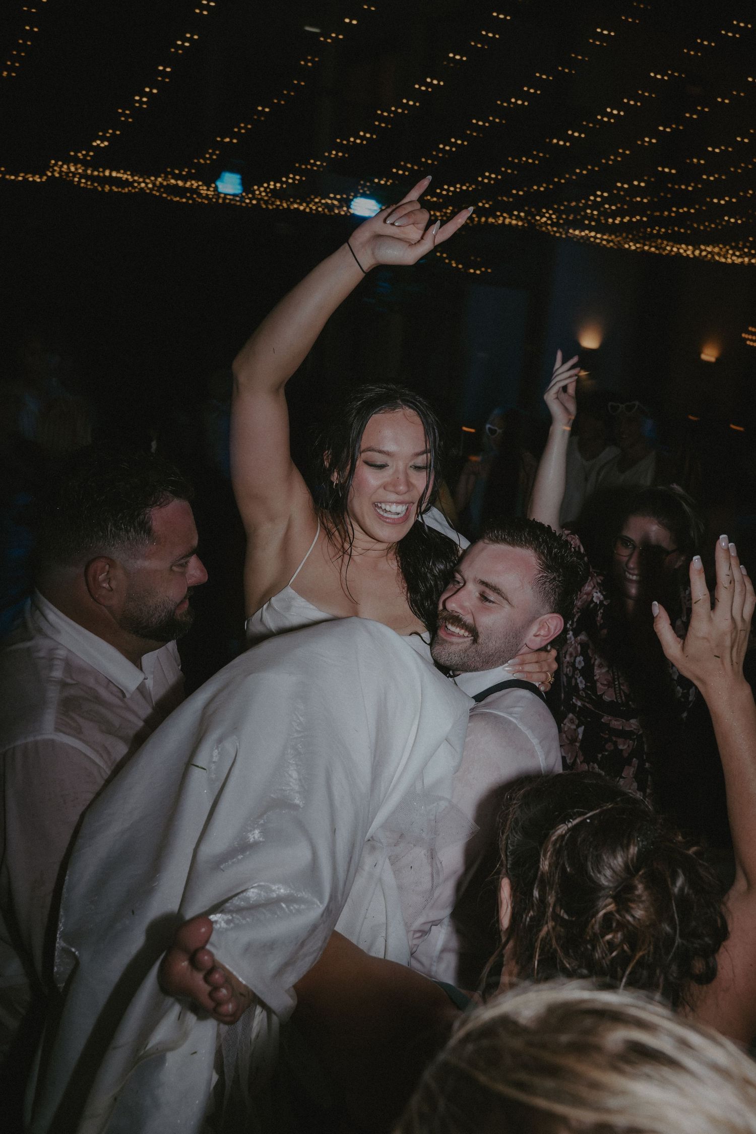 Wedding guests dance and celebrate on a dimly lit dance floor with string lights overhead.