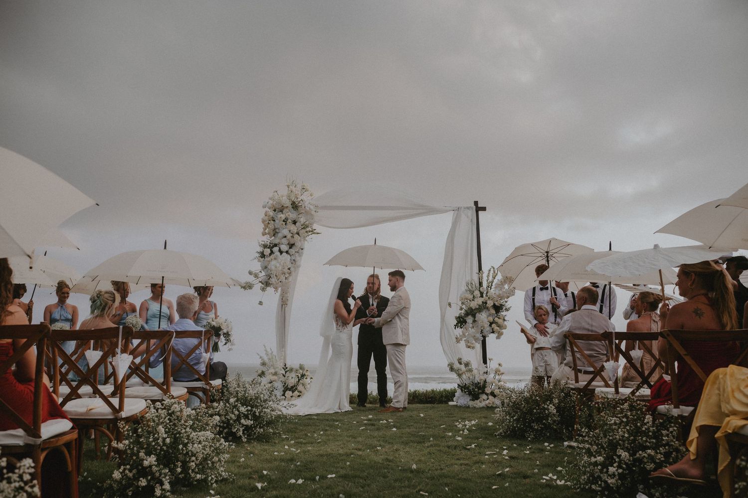 Wedding guests gather under white umbrellas on a cloudy day at an outdoor ceremony overlooking a scenic landscape.