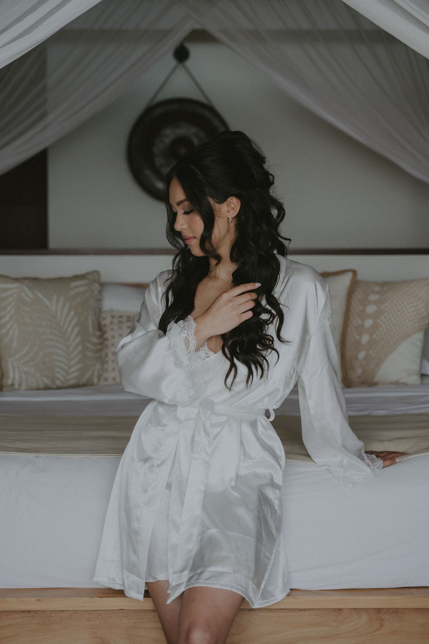 Woman in white robe sitting on bed with elegant white canopy and pillows.