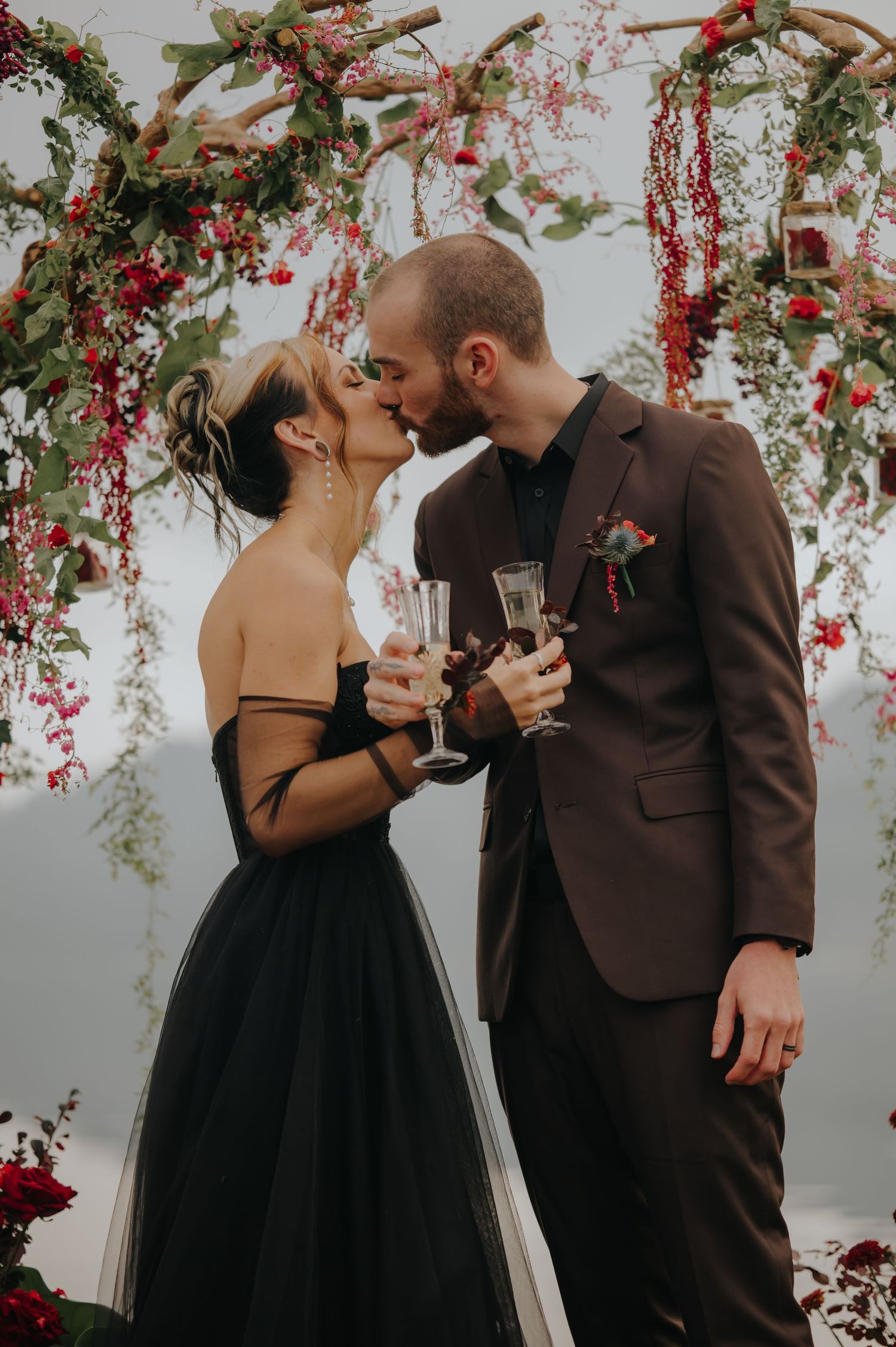 A couple shares a romantic moment under a floral arch while holding champagne glasses at their wedding celebration.