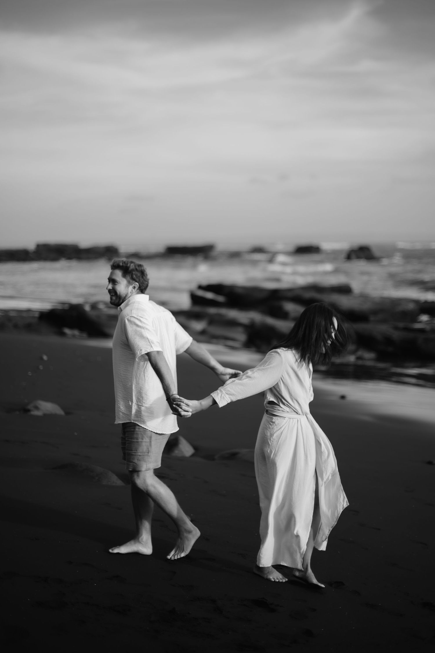 A couple in white clothing dances together on a dark beach with dramatic cloudy skies in black and white photography.