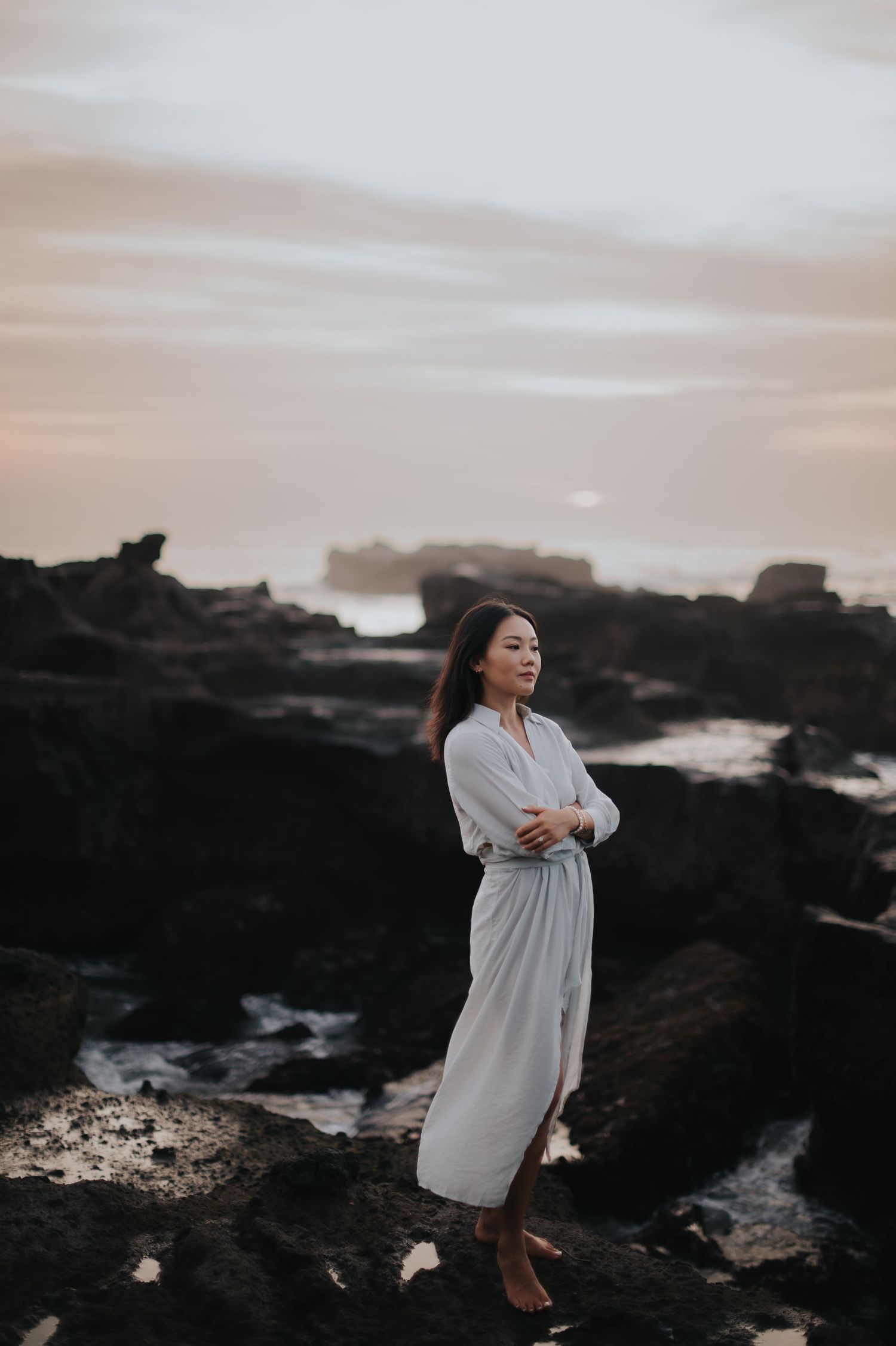 A person in a flowing white dress stands on dark rocky coastal cliffs at sunset with ocean waves crashing below.