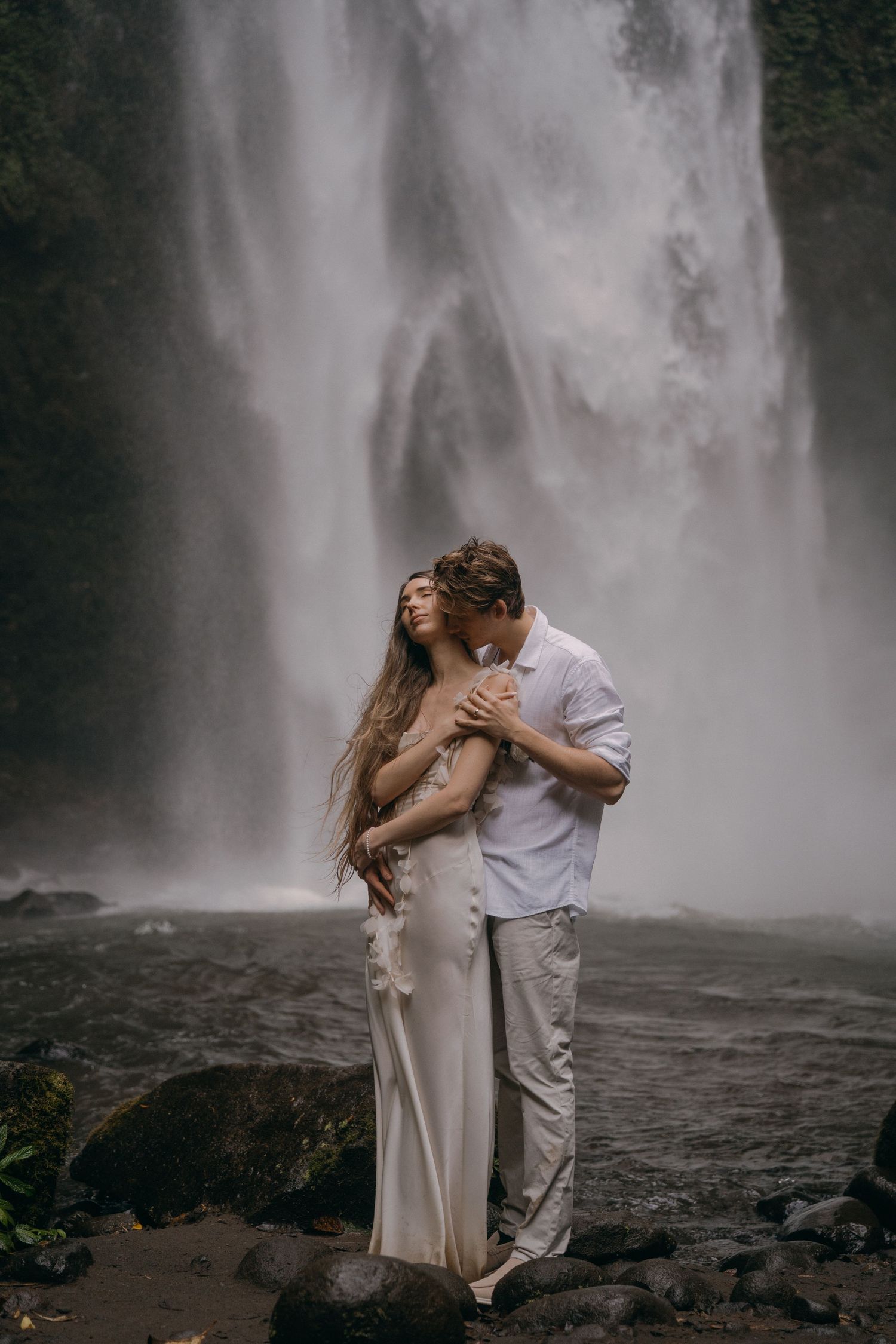 A couple shares a romantic kiss in front of a majestic waterfall cascading down dark rocky cliffs.