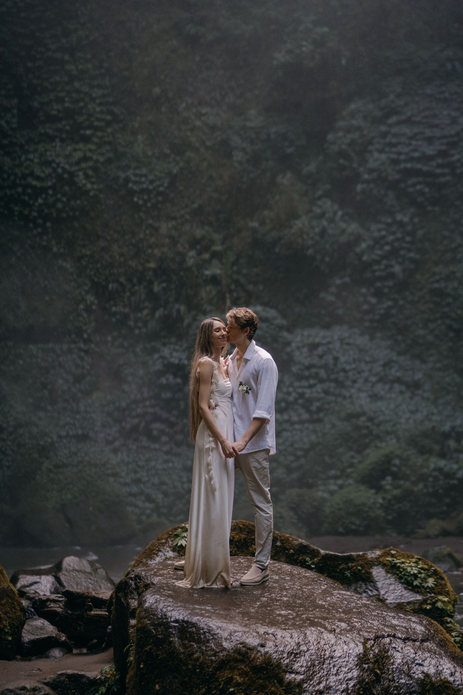 A couple shares an intimate moment while standing on a rocky cliff edge surrounded by misty mountains.