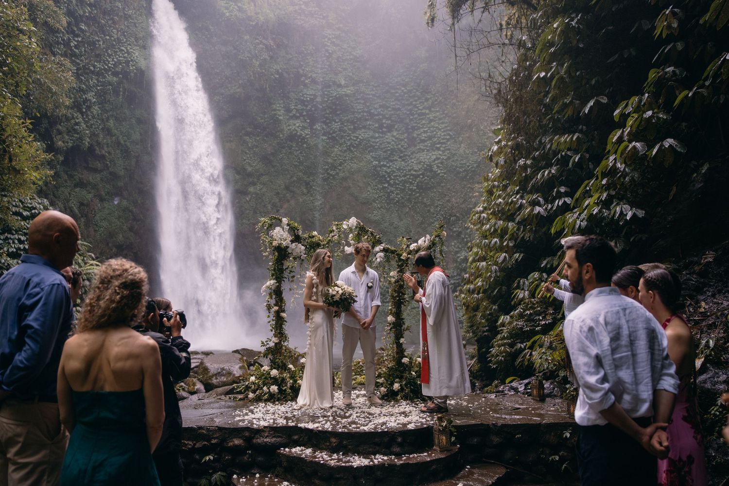 A wedding ceremony takes place in front of a majestic waterfall in a misty forest setting.