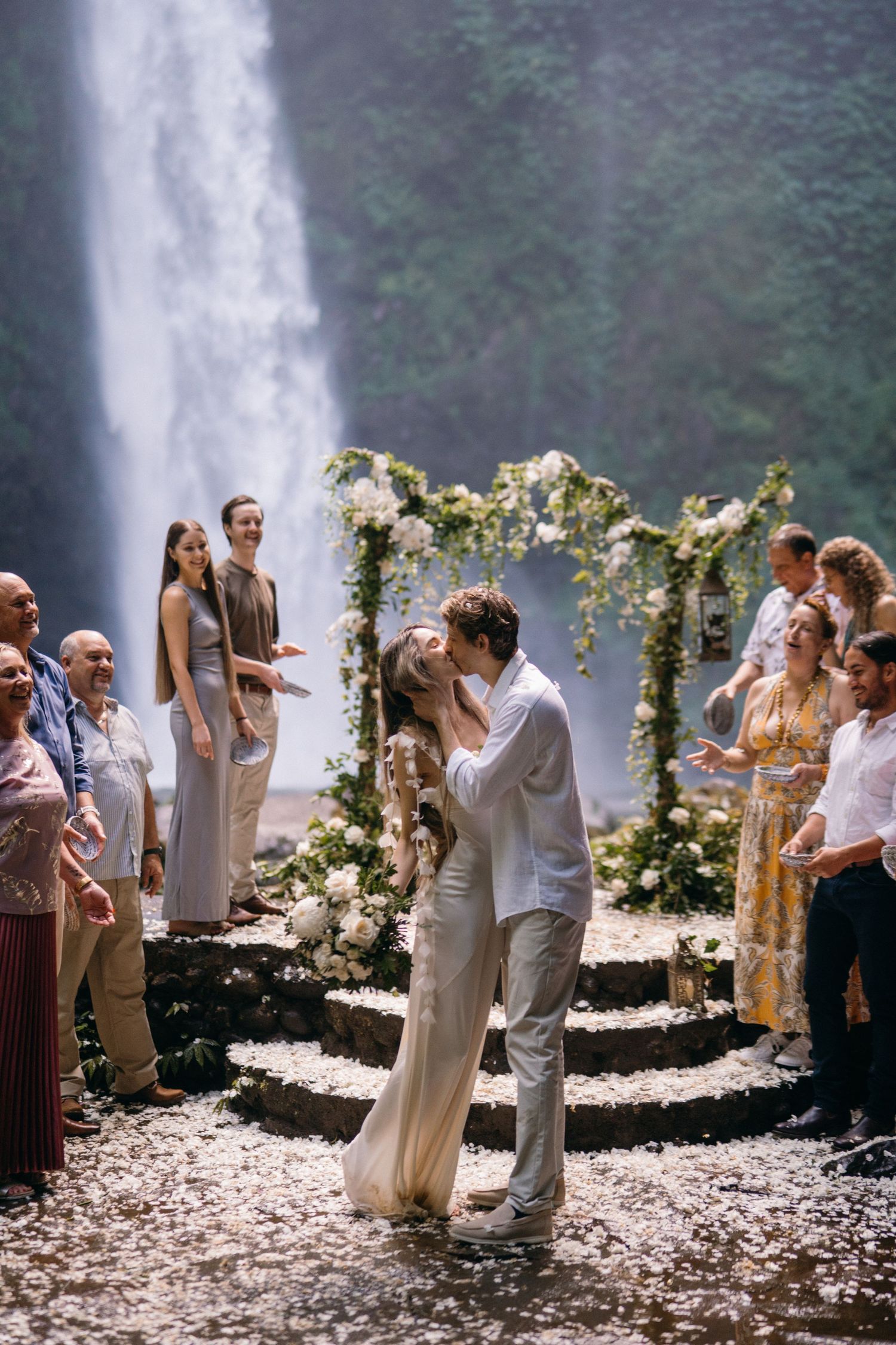A wedding ceremony takes place in front of a majestic waterfall with a floral arch and guests watching from stone steps.