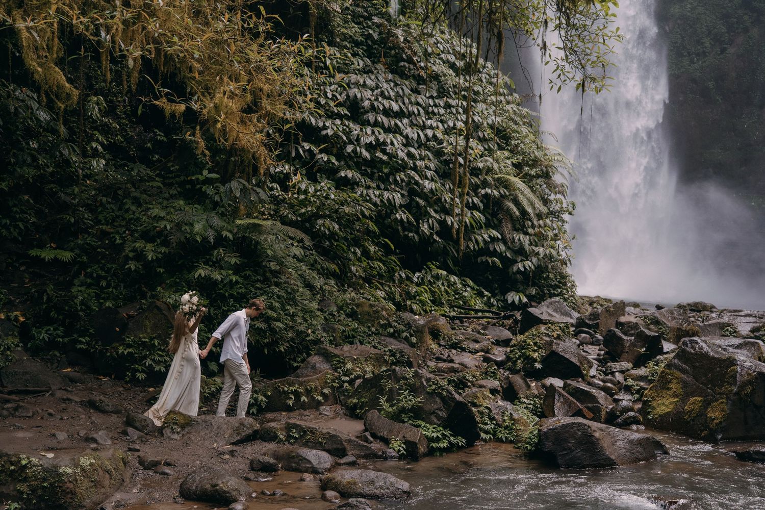 Two people in formal attire walk along rocky path near majestic waterfall surrounded by lush greenery.