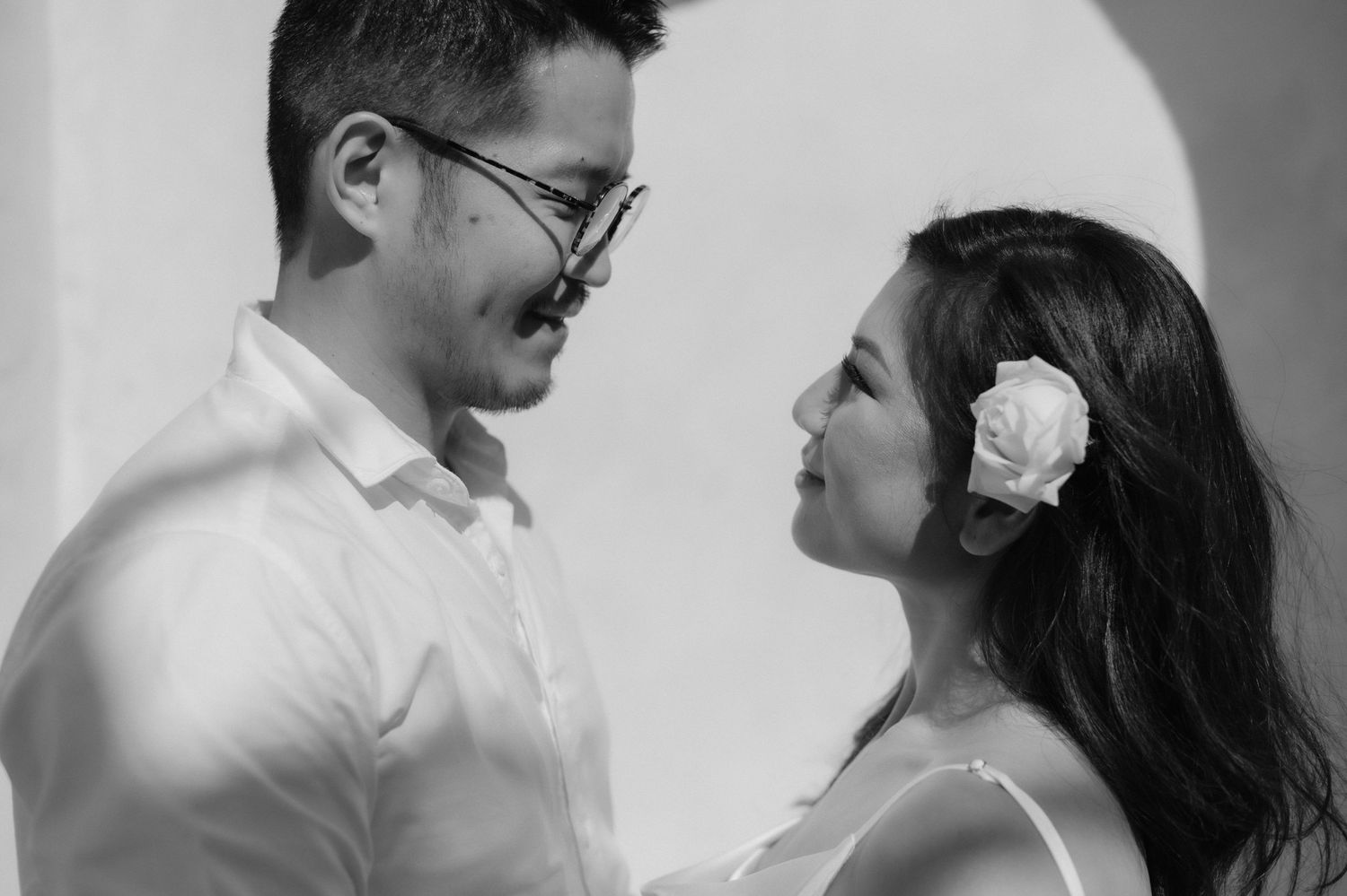 Black and white photo of a couple in romantic pose, man wearing glasses, woman with white flower in hair.