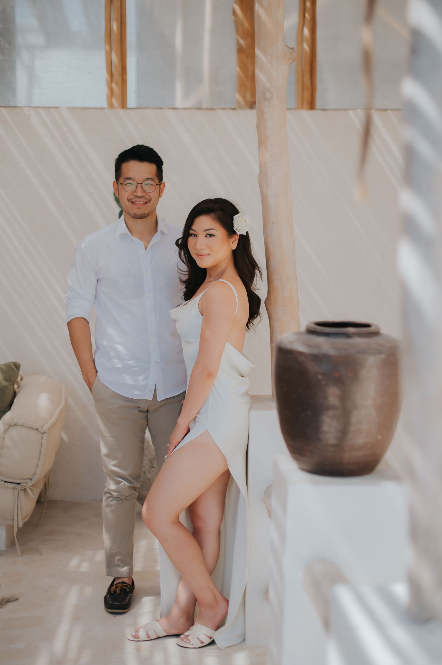 A couple poses elegantly in a bright, airy space with white drapes and a rustic ceramic vase nearby.