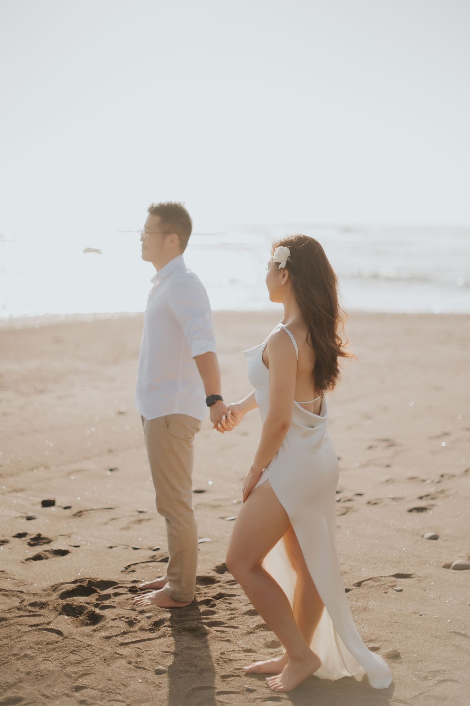 Couple holding hands on a sandy beach at sunset, woman in white dress with sunglasses, man in white shirt and khaki pants.