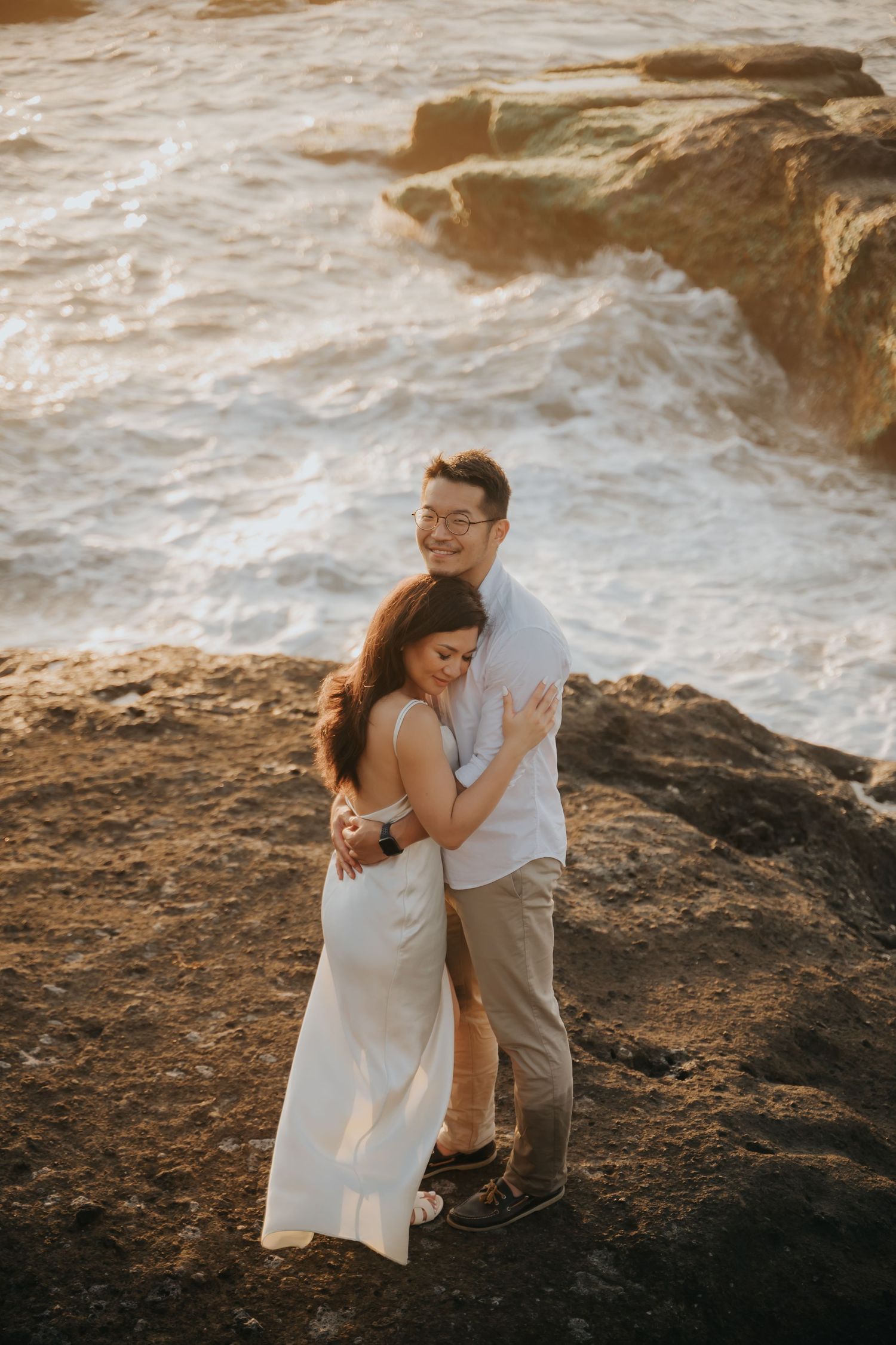 Couple embracing on rocky coastal cliffs at golden hour, waves crashing behind them, woman in white dress, man in casual attire.