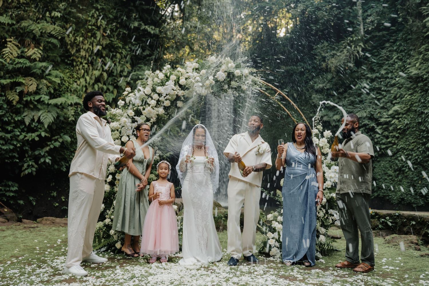 Wedding party poses before a white floral arch and waterfall backdrop in tropical Bali setting.