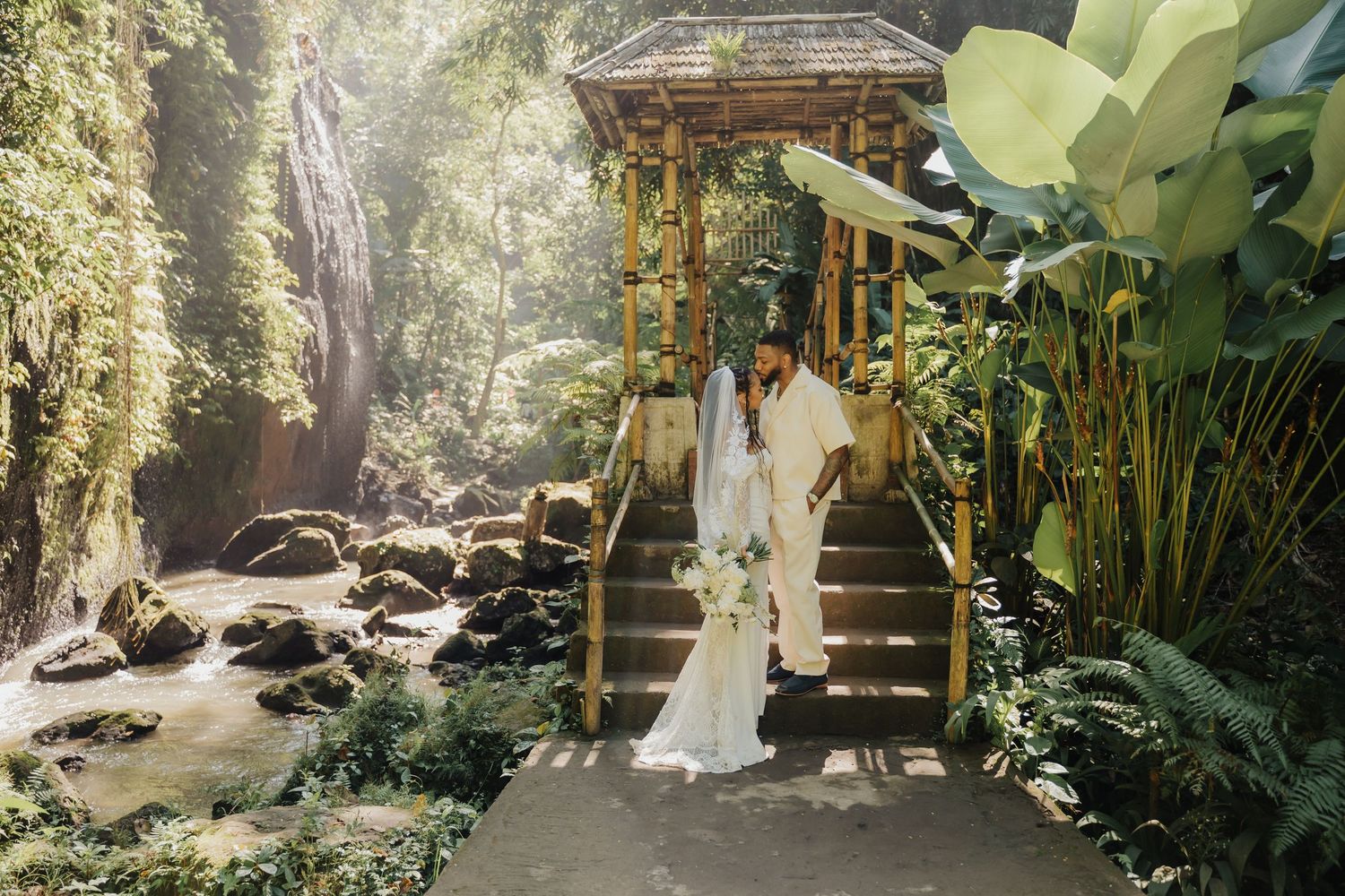 Bride and groom posing on bamboo steps surrounded by tropical greenery and waterfall in Bali.