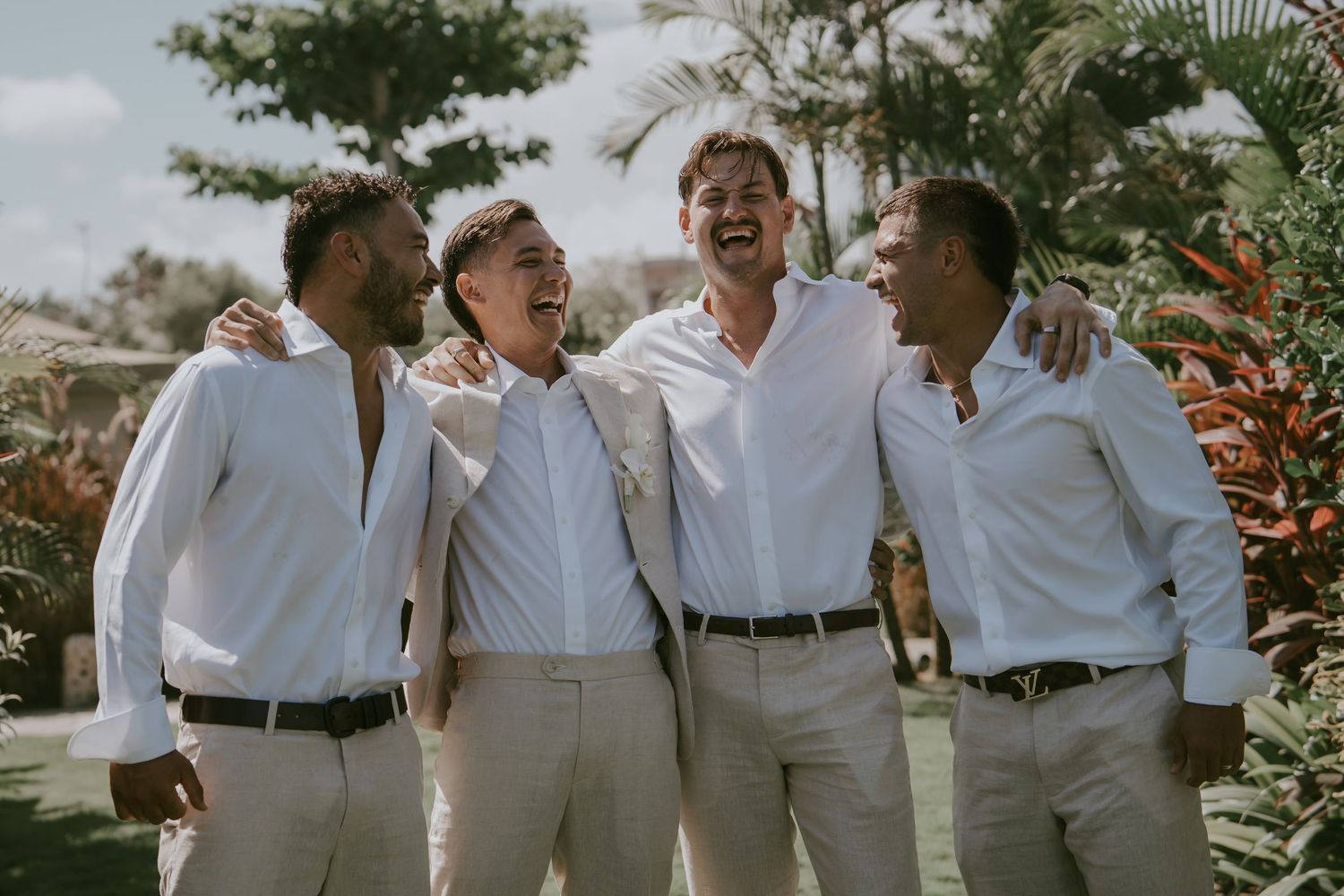 A group wearing matching white outfits laughs together in a tropical garden setting during a photo session.
