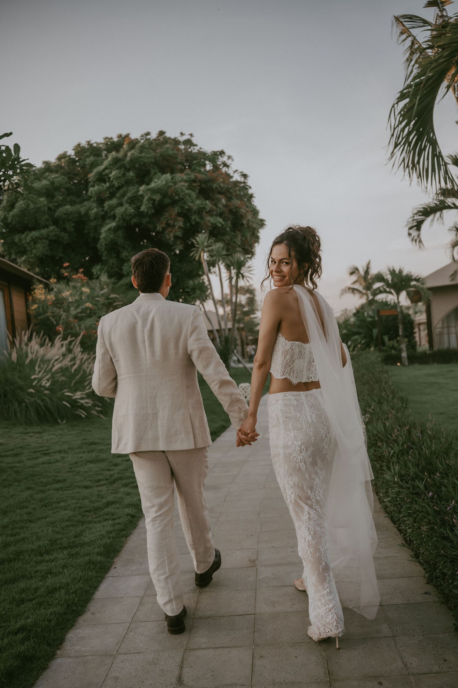 Newlywed couple in white attire walking hand in hand down a palm-lined path at sunset.