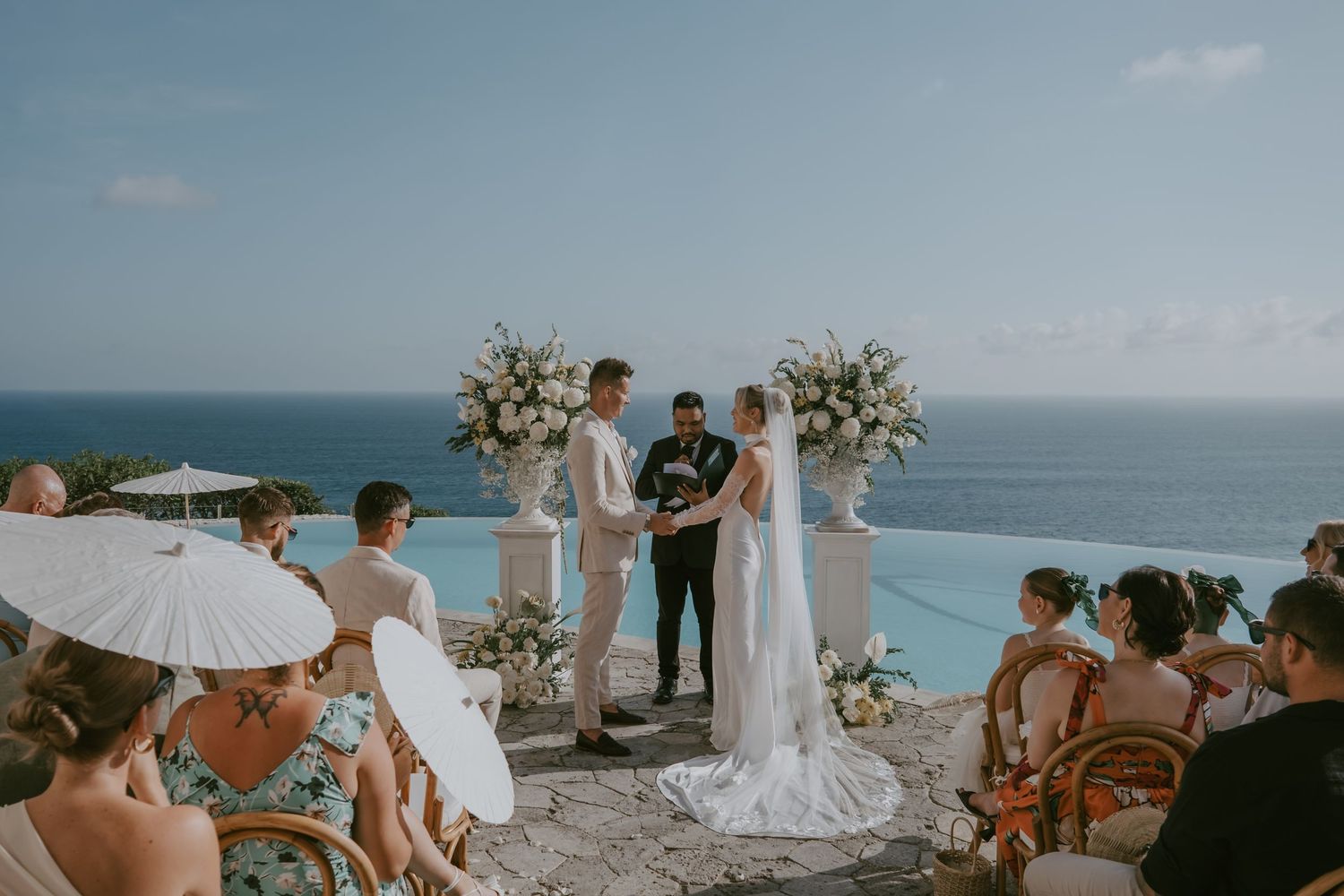 Beachside wedding ceremony setup with white floral arrangements against ocean backdrop.