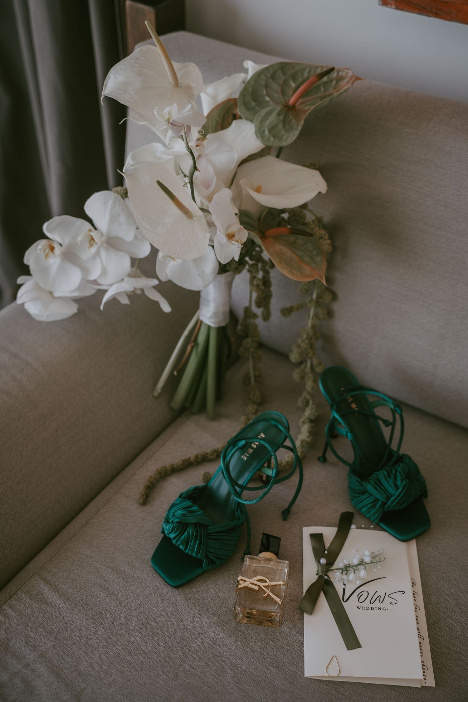 Wedding details featuring white orchids, emerald green shoes and invitation cards on neutral surface.