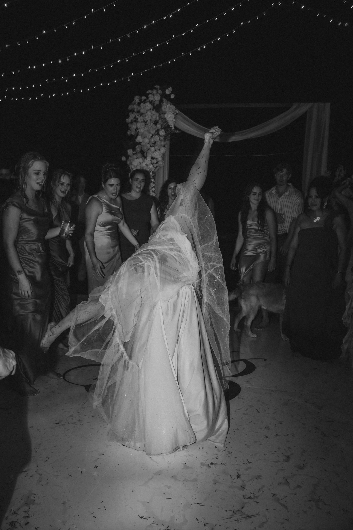 A bride in a white wedding dress dances with guests under twinkling lights at an evening reception celebration.