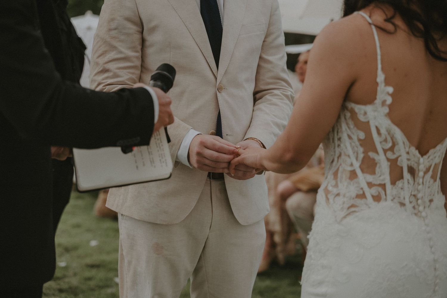 A couple in wedding attire holds hands during their outdoor ceremony, with the bride in white and groom in beige.