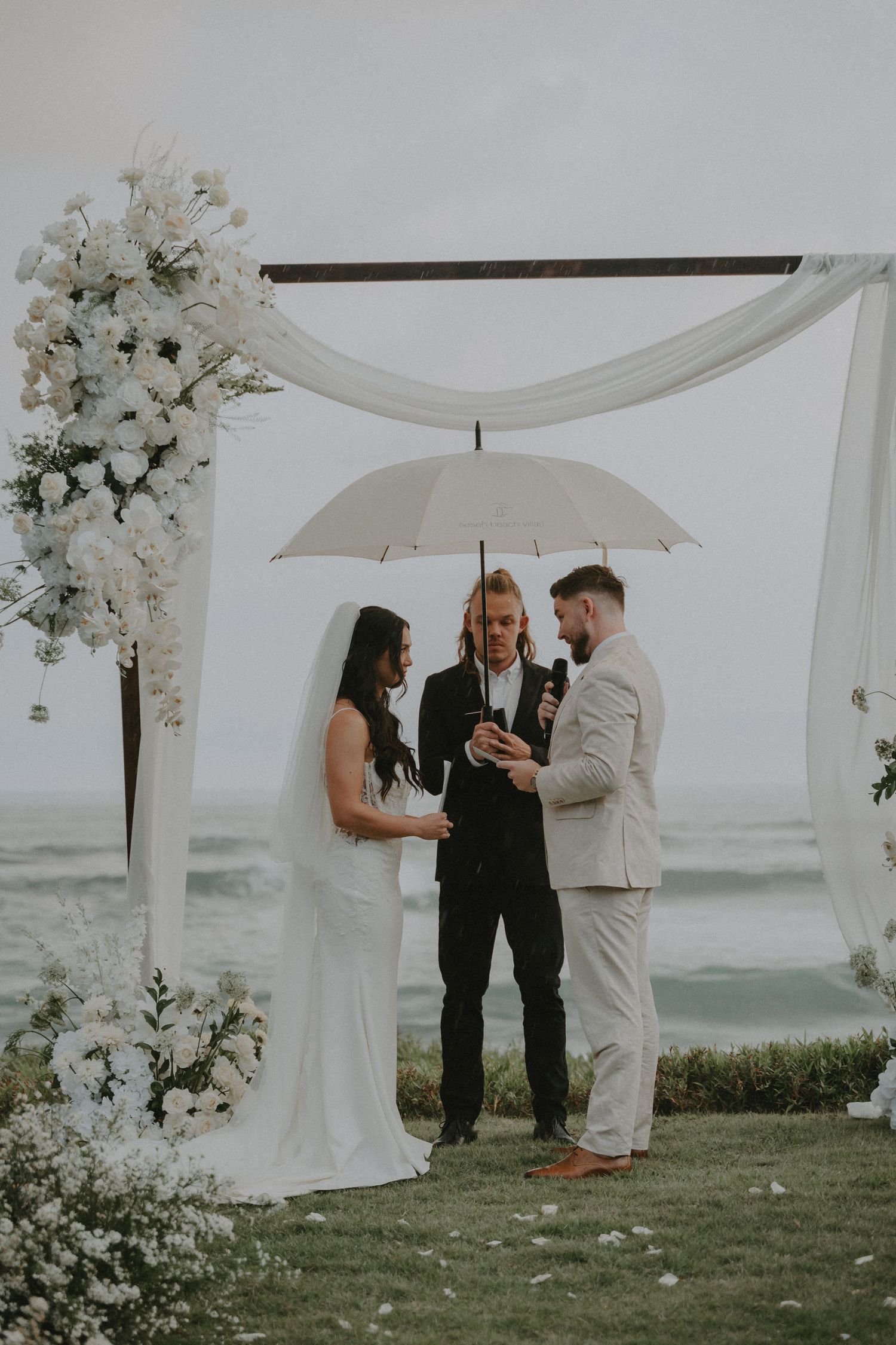 A wedding ceremony takes place on a misty oceanfront lawn with white floral arrangements and an elegant white canopy.