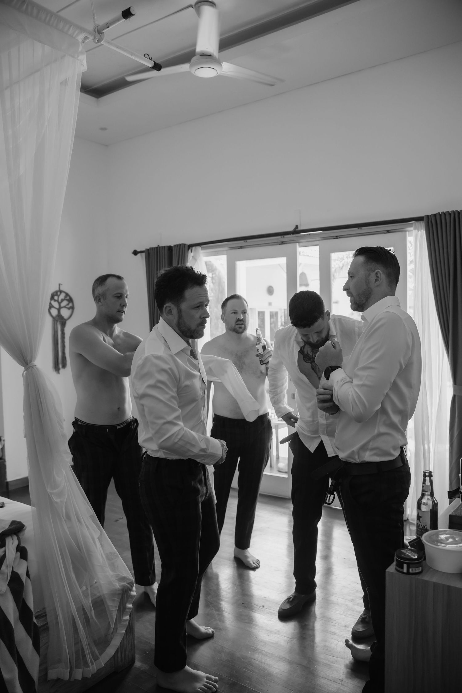 Black and white photo of groomsmen getting ready in a bright room with large windows.