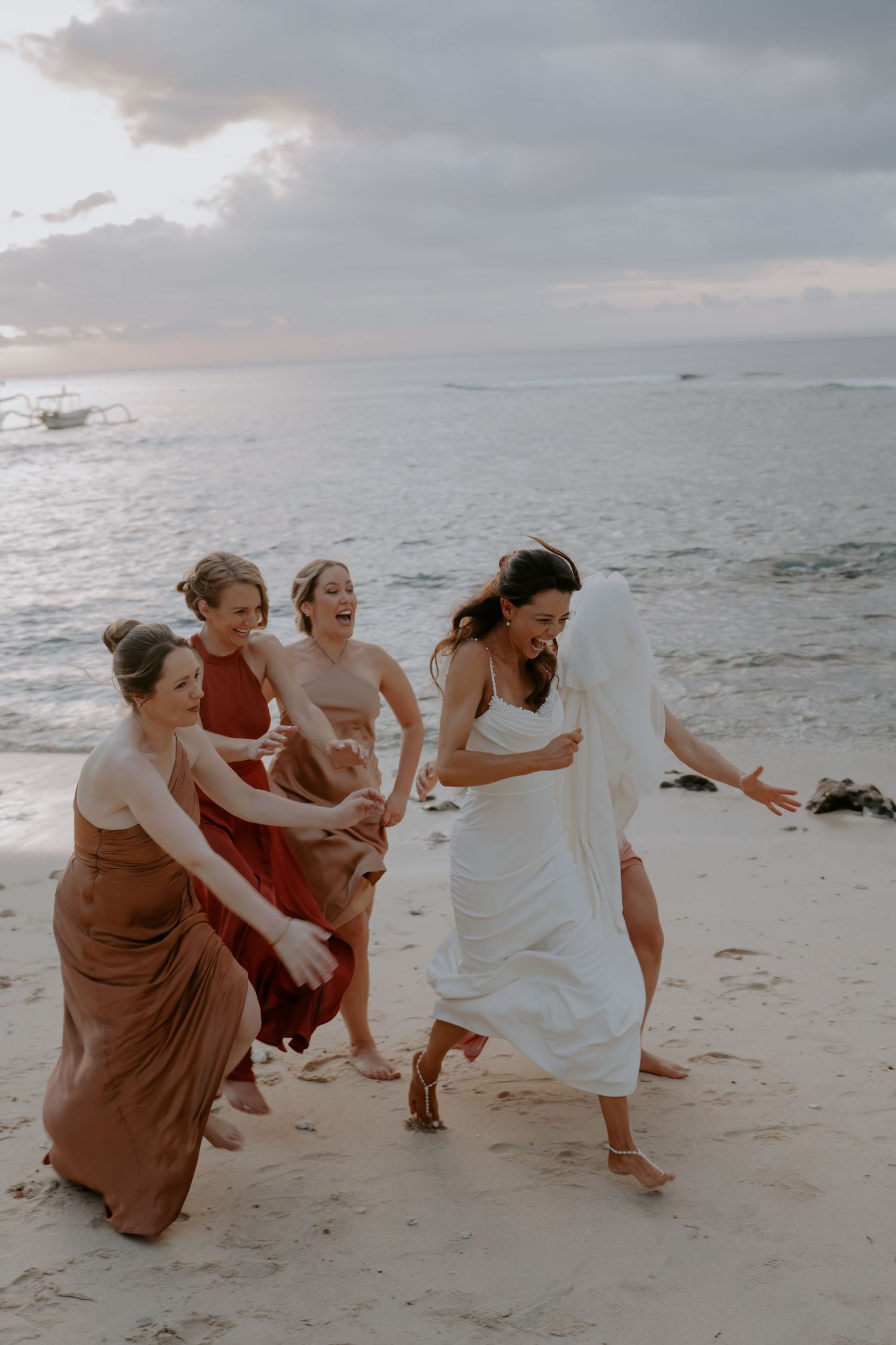 A group in flowing dresses playfully poses together on a cloudy beach at sunset.
