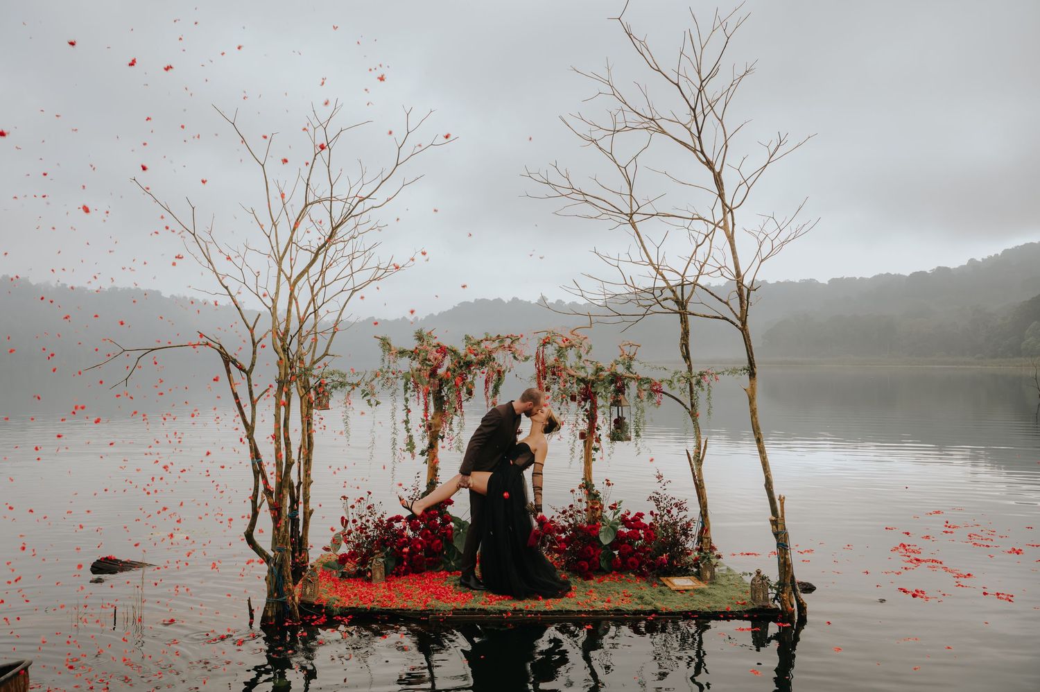 Romantic ceremony setup on floating dock with red flower petals scattered in misty lake setting.