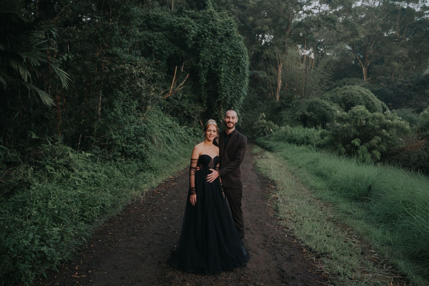A couple in dark attire walks along a scenic wooded path during a romantic photoshoot.