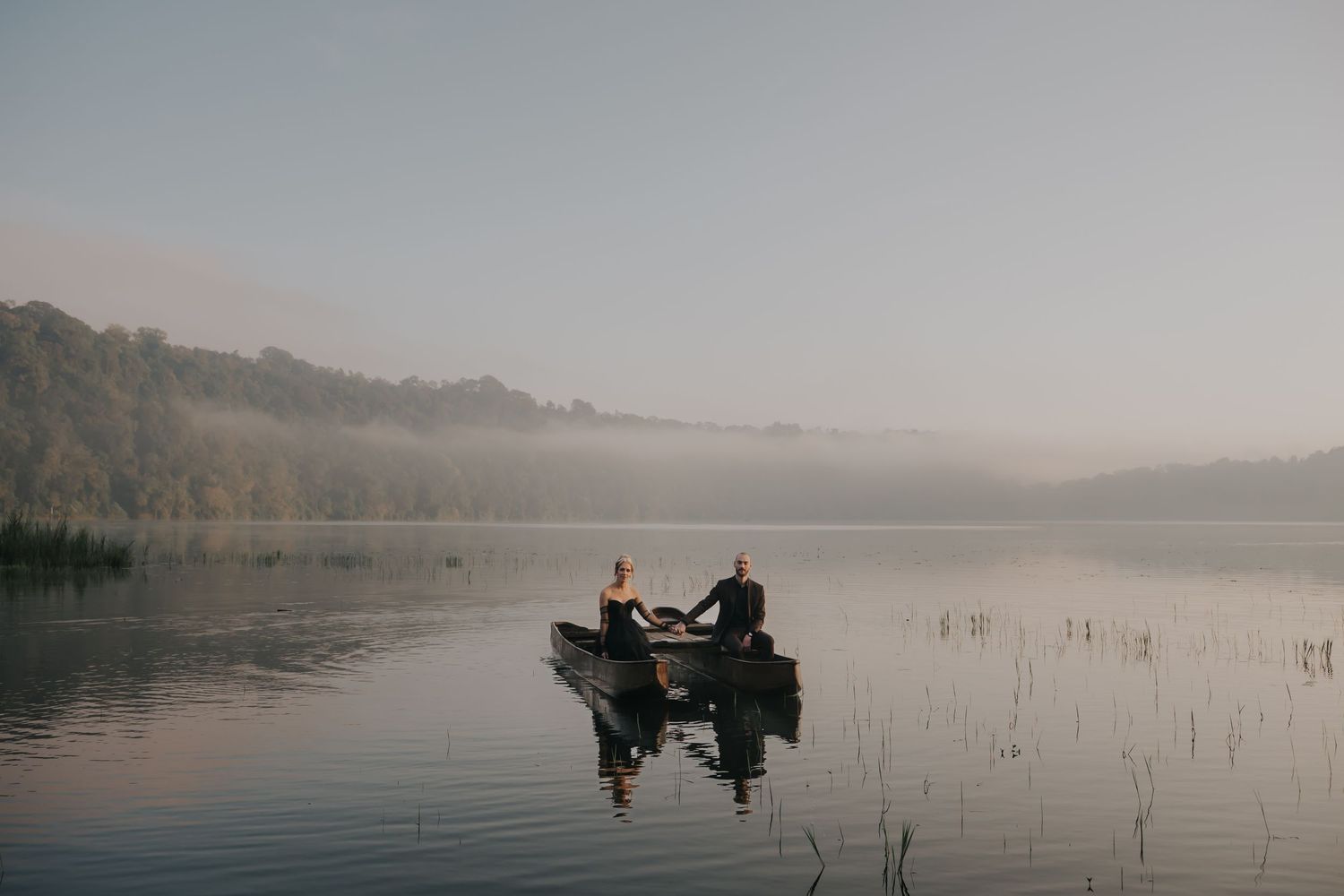 Couple paddling wooden canoes across a misty lake at sunrise with trees reflecting in calm waters.