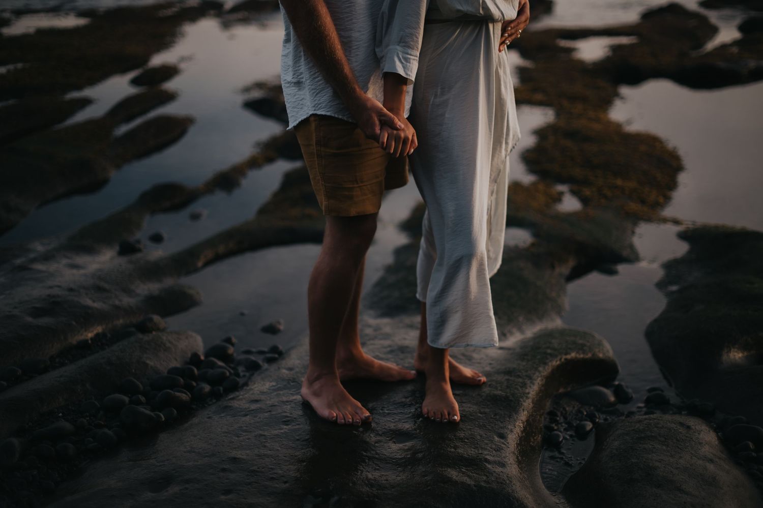 Couple's feet standing on rocky beach during sunset with water reflections creating romantic silhouette.