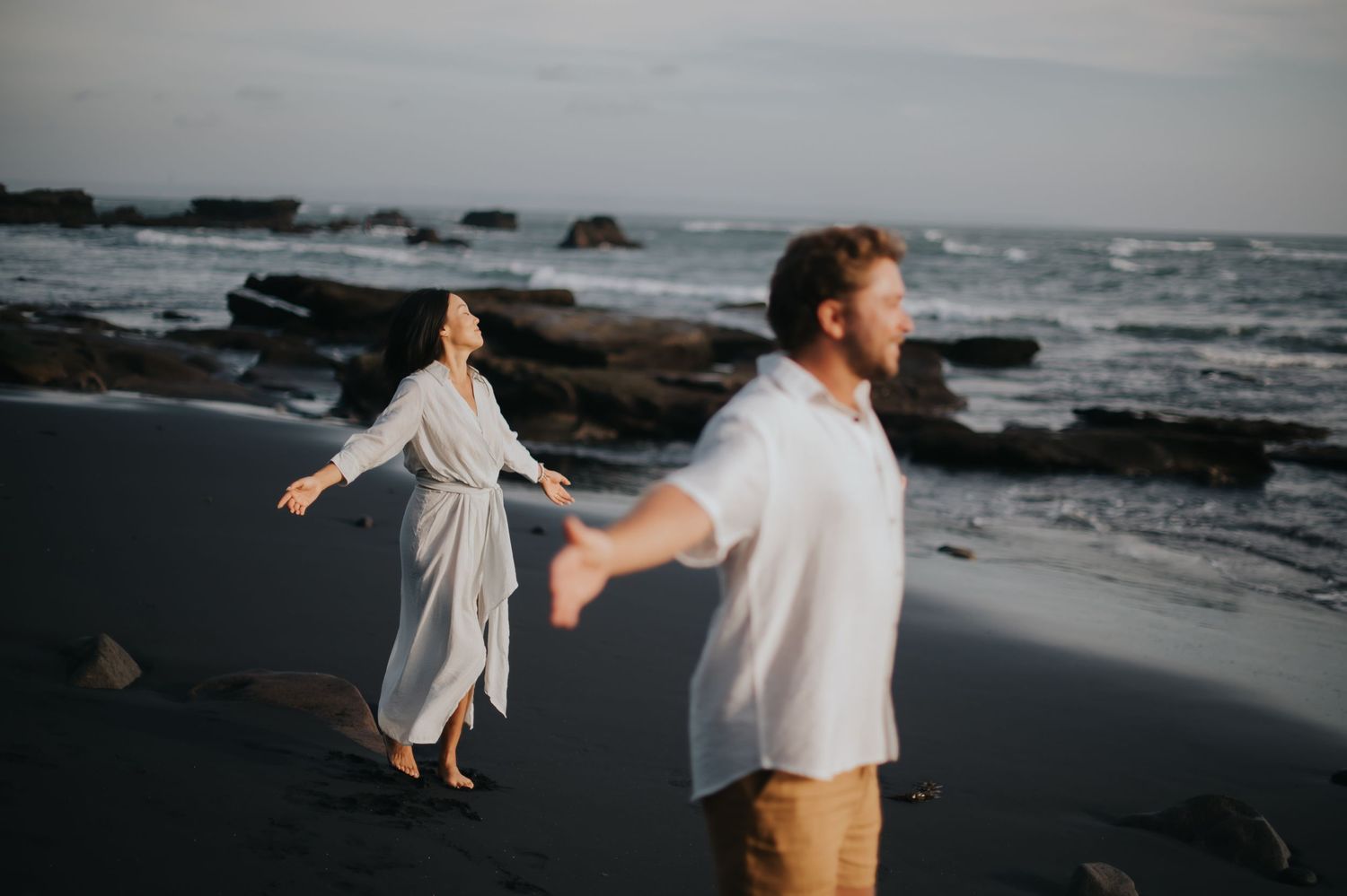 Two people in white clothing dance together on a rocky beach at dusk with ocean waves crashing in the background.