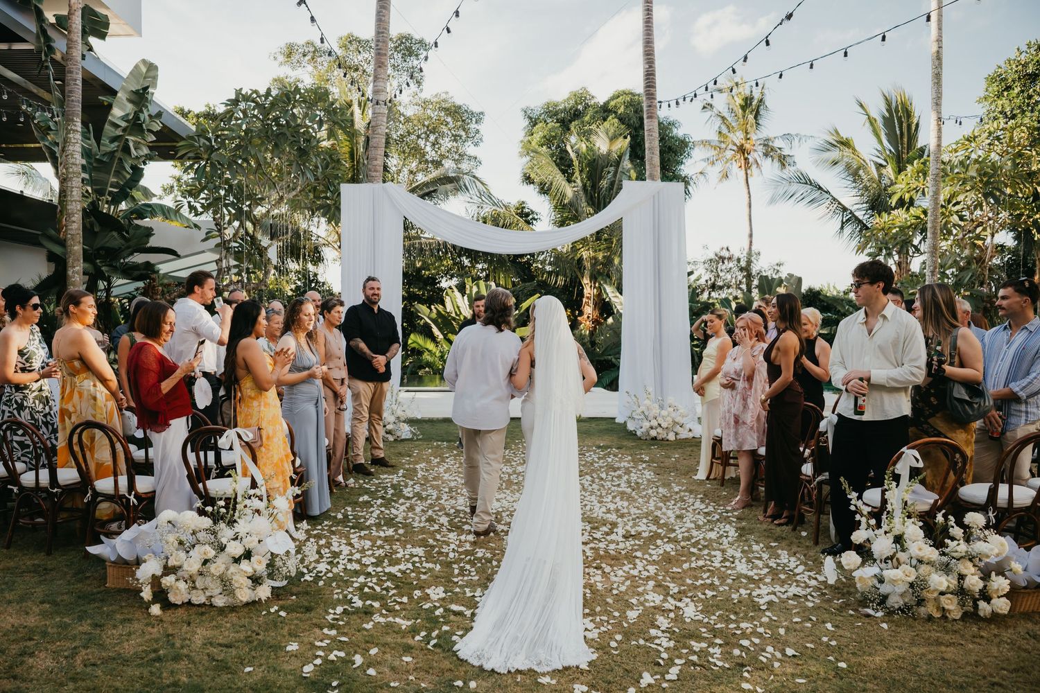 Bride approaching groom at outdoor wedding altar surrounded by white flowers and wedding guests.