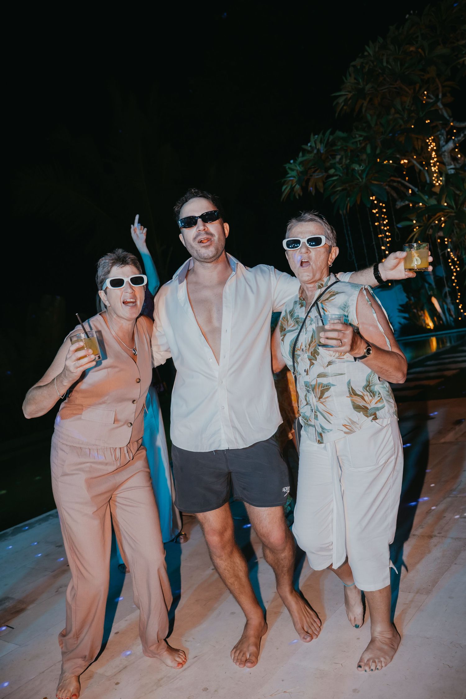 Three guests in sunglasses pose and celebrate poolside at an outdoor evening wedding reception.