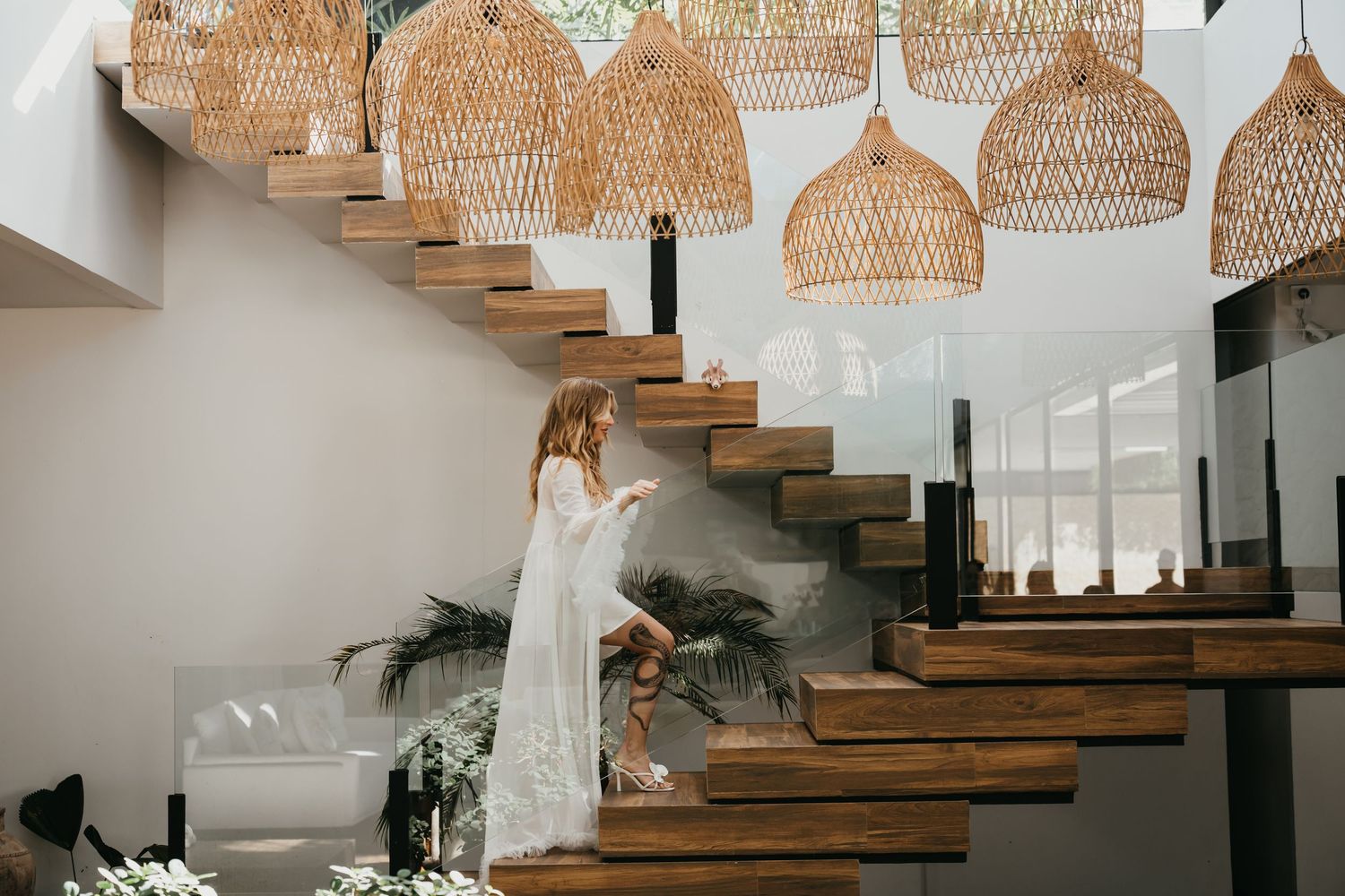 Woman in white lace dress descending wooden floating stairs under woven rattan pendant lights in modern boho interior.