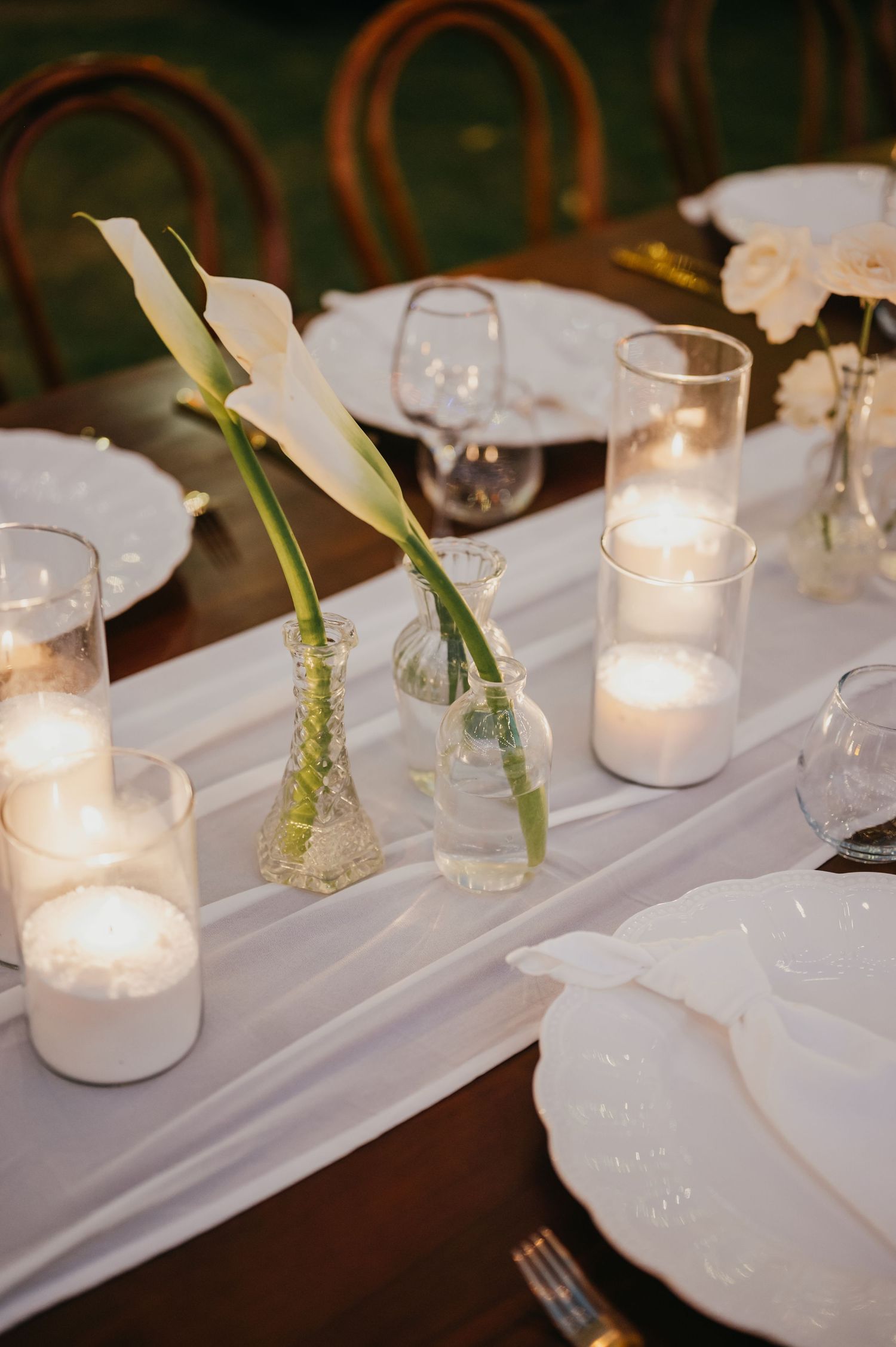 Close-up of wedding table centerpiece featuring white calla lilies in glass bud vases with glowing pillar candles.