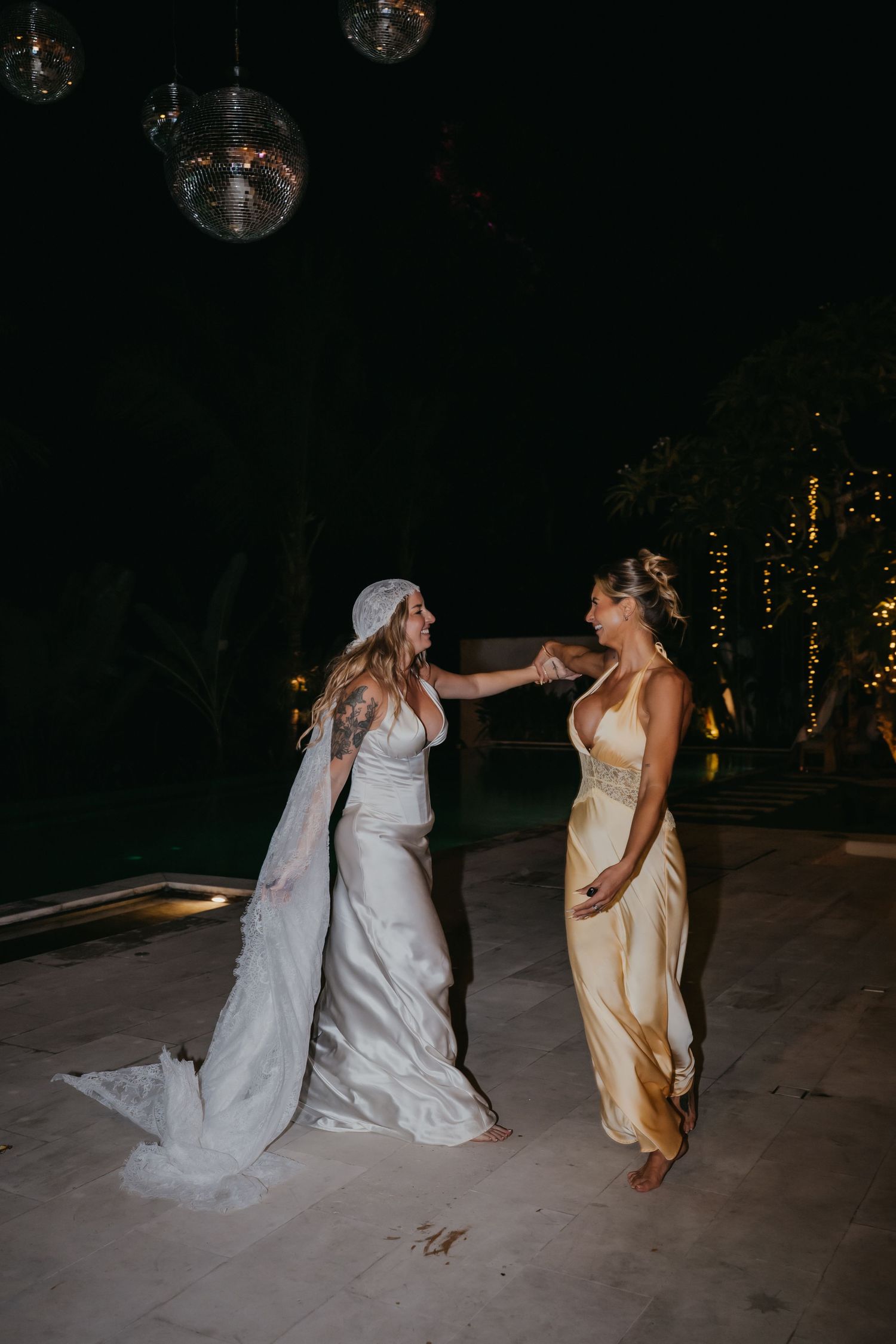 Bride in white satin gown and veil dances with bridesmaid in gold dress under disco balls at night.