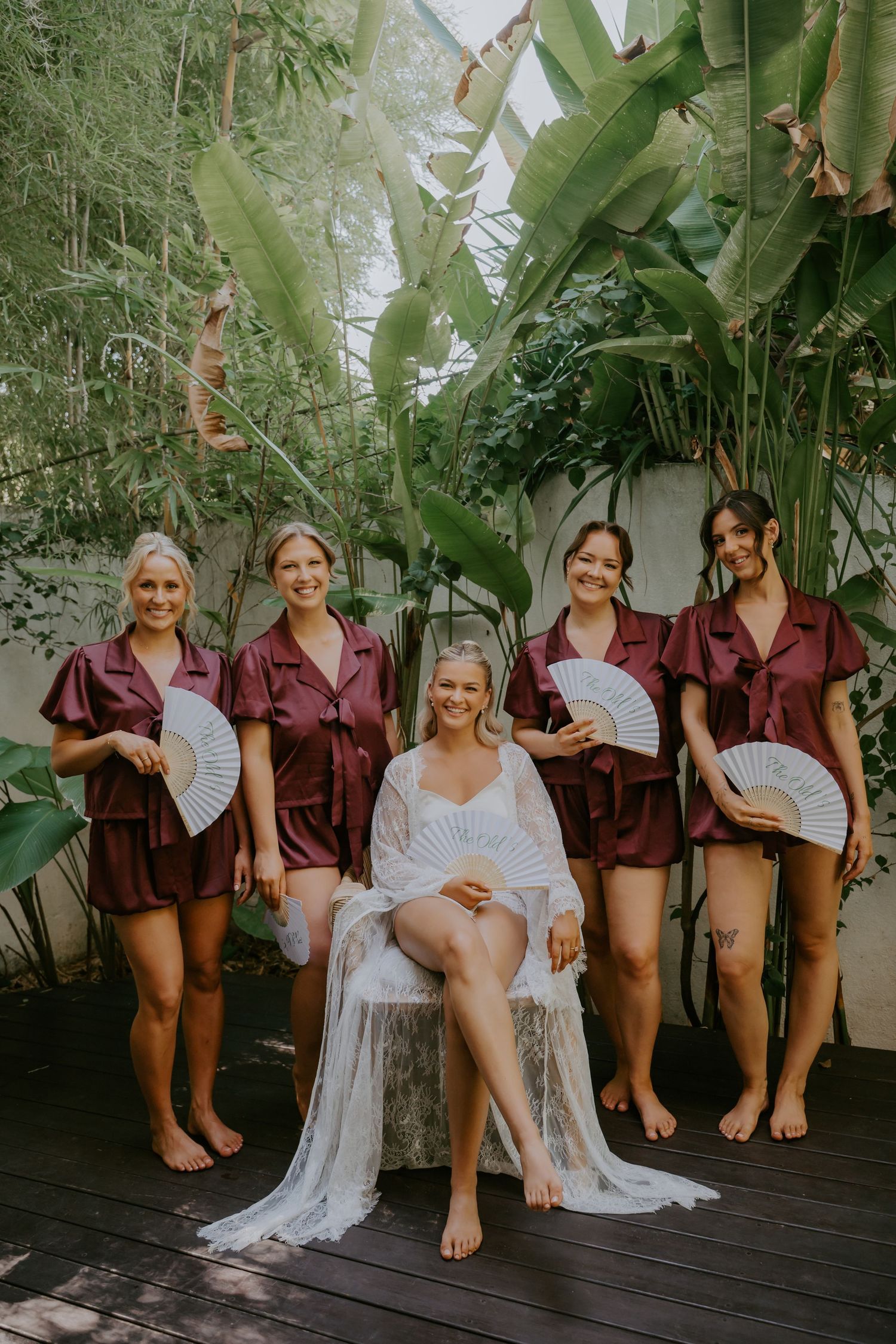 Bride in white robe sits with four bridesmaids in burgundy satin sets holding fans amid tropical greenery.