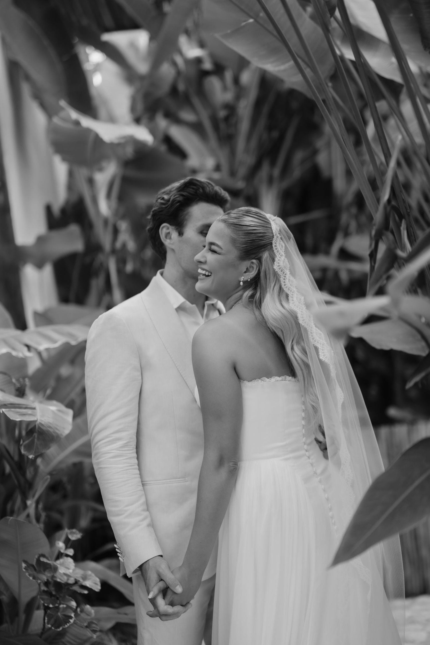 Black and white portrait of smiling bride and groom holding hands among tropical garden greenery.