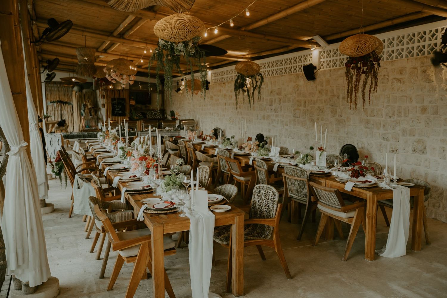 Long wooden banquet tables elegantly set for tropical wedding reception in open-air venue with string lights.