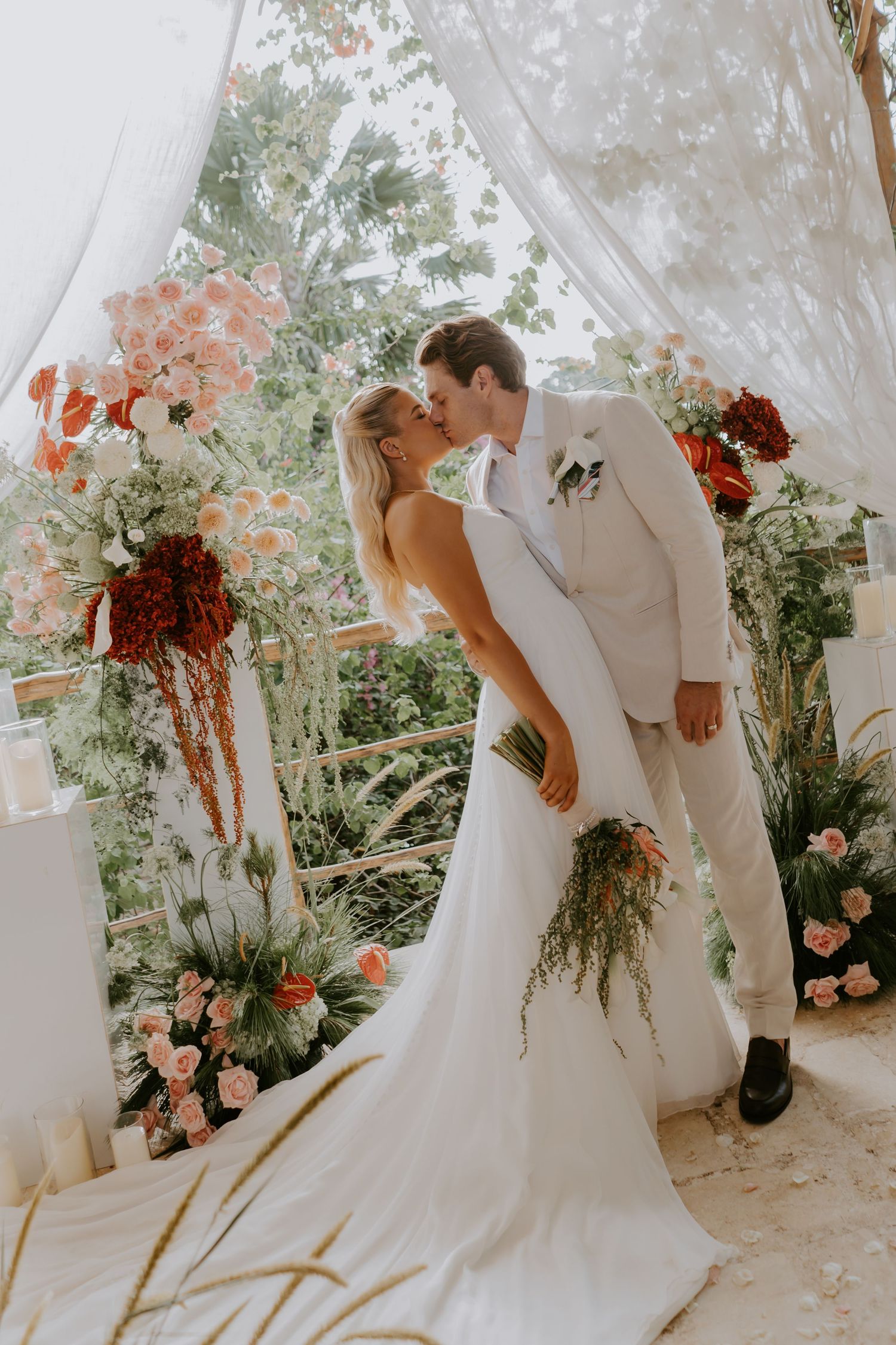 Bride and groom sharing first kiss at floral altar with red and pink blooms.