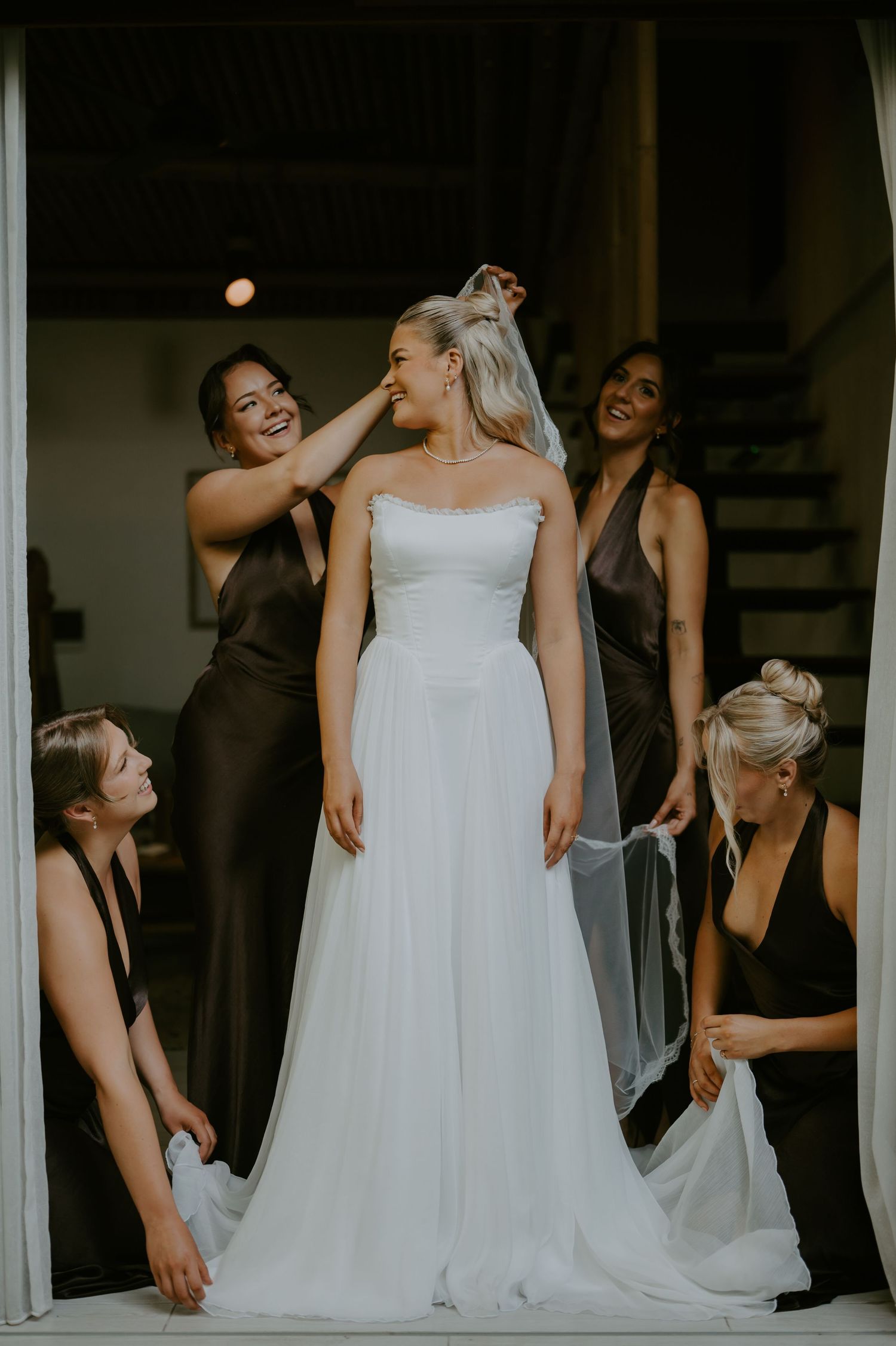 Bridesmaids in dark gowns help smiling bride into her strapless white wedding dress in a doorway.