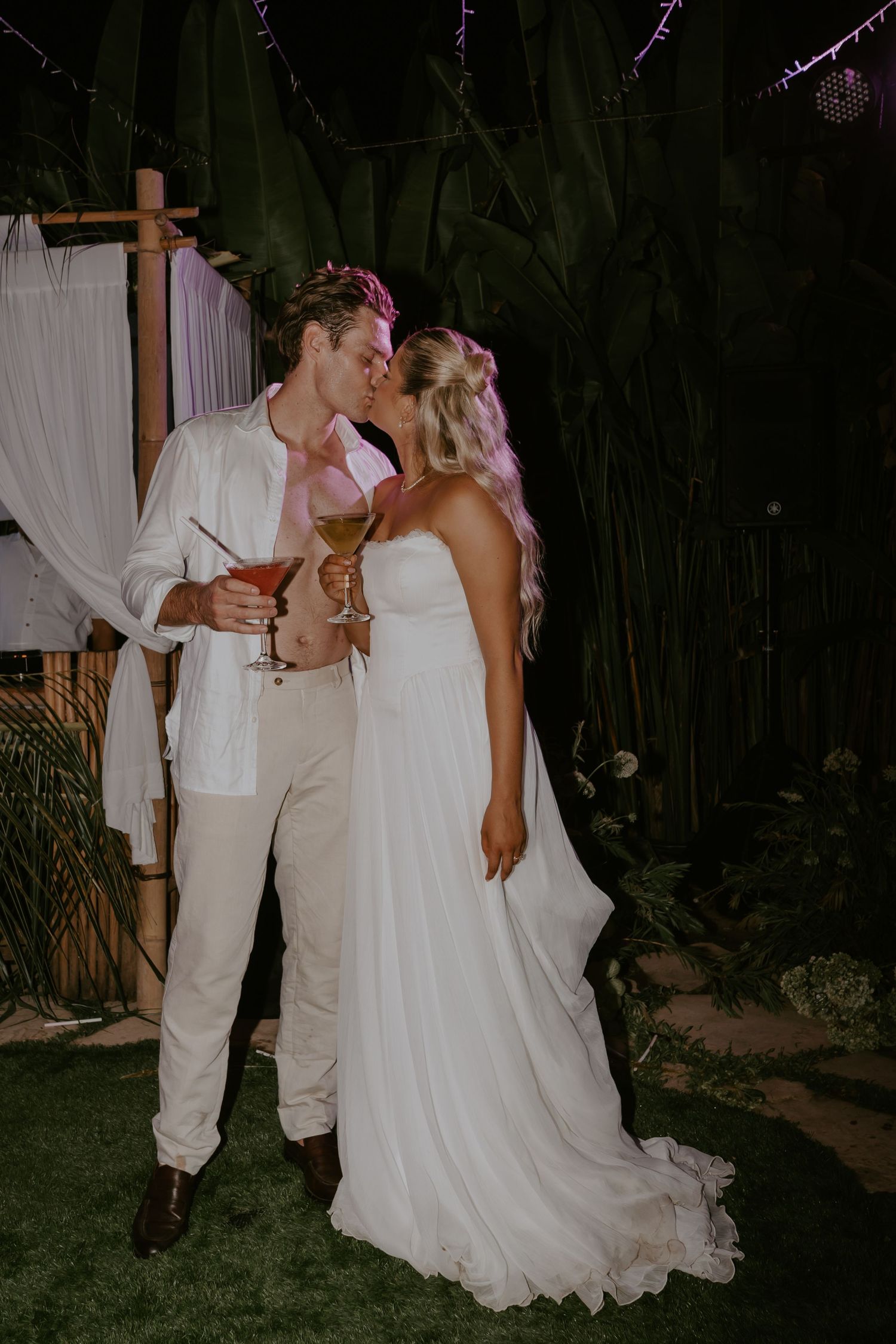 Bride and groom share a kiss while holding cocktails at outdoor evening reception.